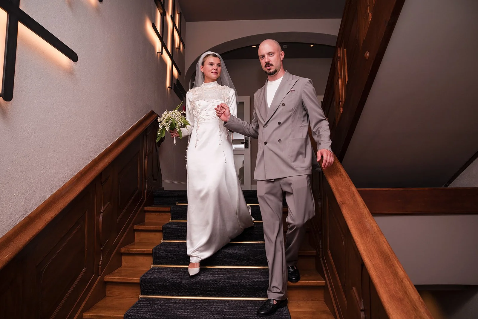 Ein Brautpaar geht die Treppe in einem Haus hinunter. Die Braut trägt ein langes weißes Brautkleid mit Schleier und hält einen Blumenstrauß. Der Bräutigam trägt einen grauen Anzug. Sie sind im Flur mit Holzwandverkleidung und moderner Beleuchtung.
