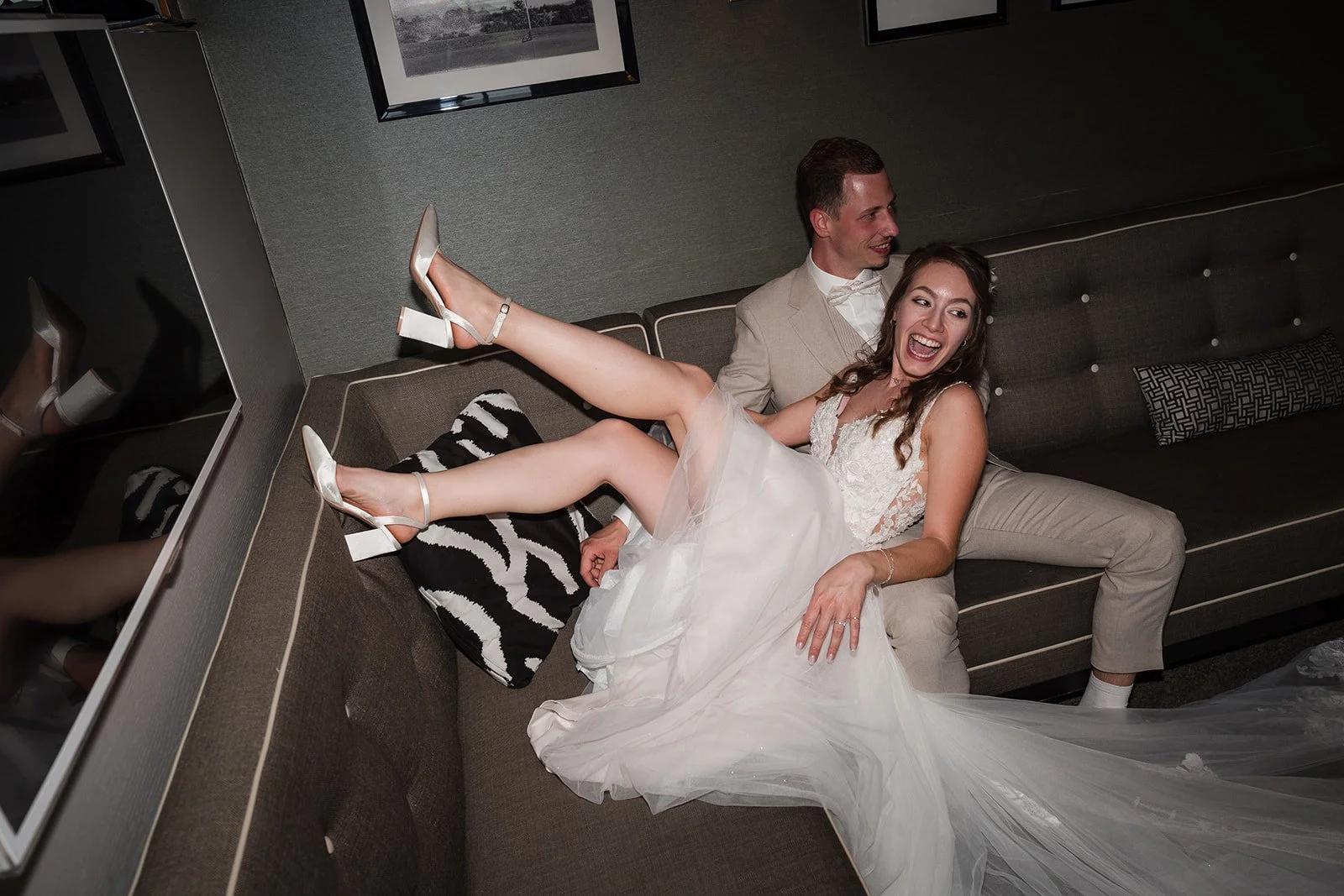 Ein glückliches Brautpaar beim Hochzeitsfoto, die Braut trägt ein weißes Kleid und die Schuhe mit hohen Absätzen, der Bräutigam in einem beige Anzug, sie sitzen auf einer schwarzen Couch mit dunklem Hintergrund.