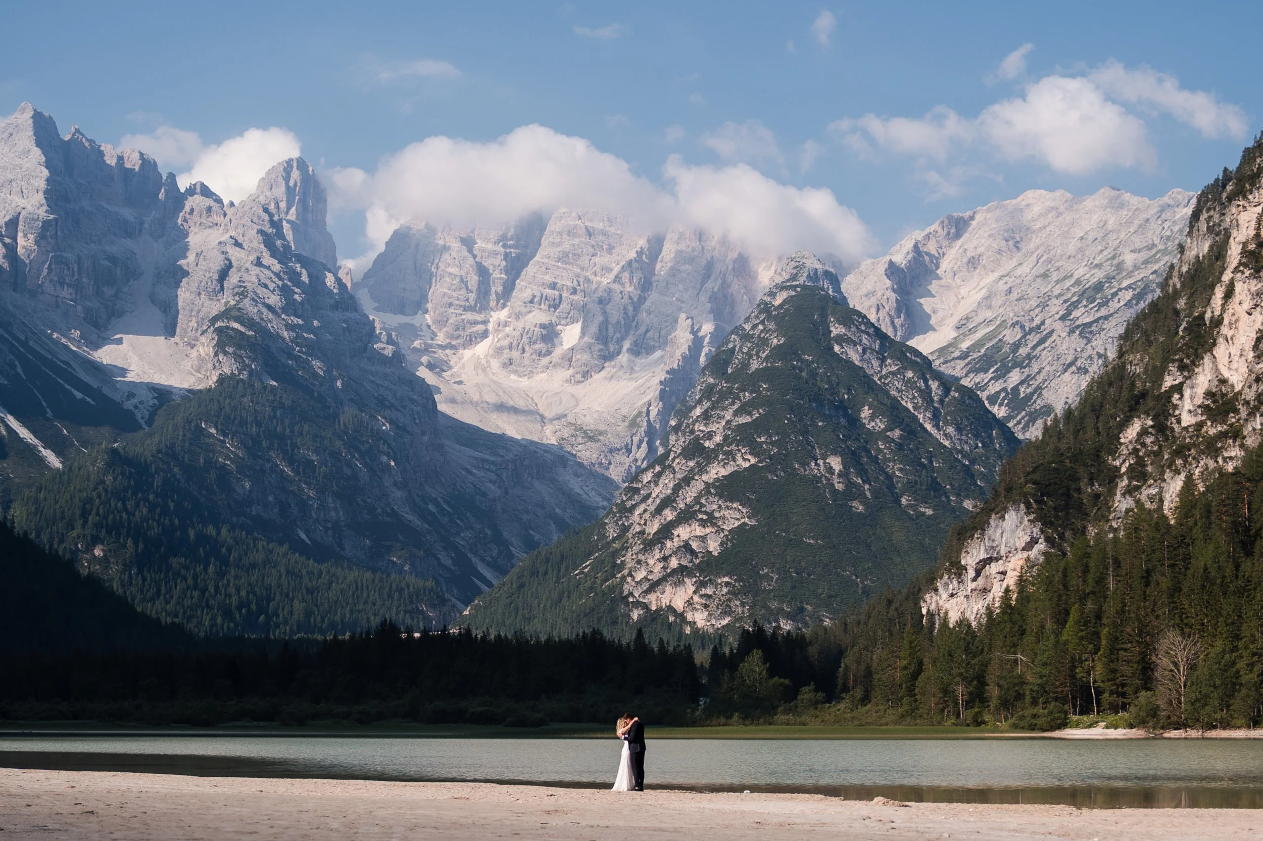 Ein Paar steht am Ufer eines Sees mit einer Berglandschaft im Hintergrund, umgeben von hohen Bergen mit Schneespitzen, einige Wolken am Himmel, ruhiges Wasser, romantische Atmosphäre.