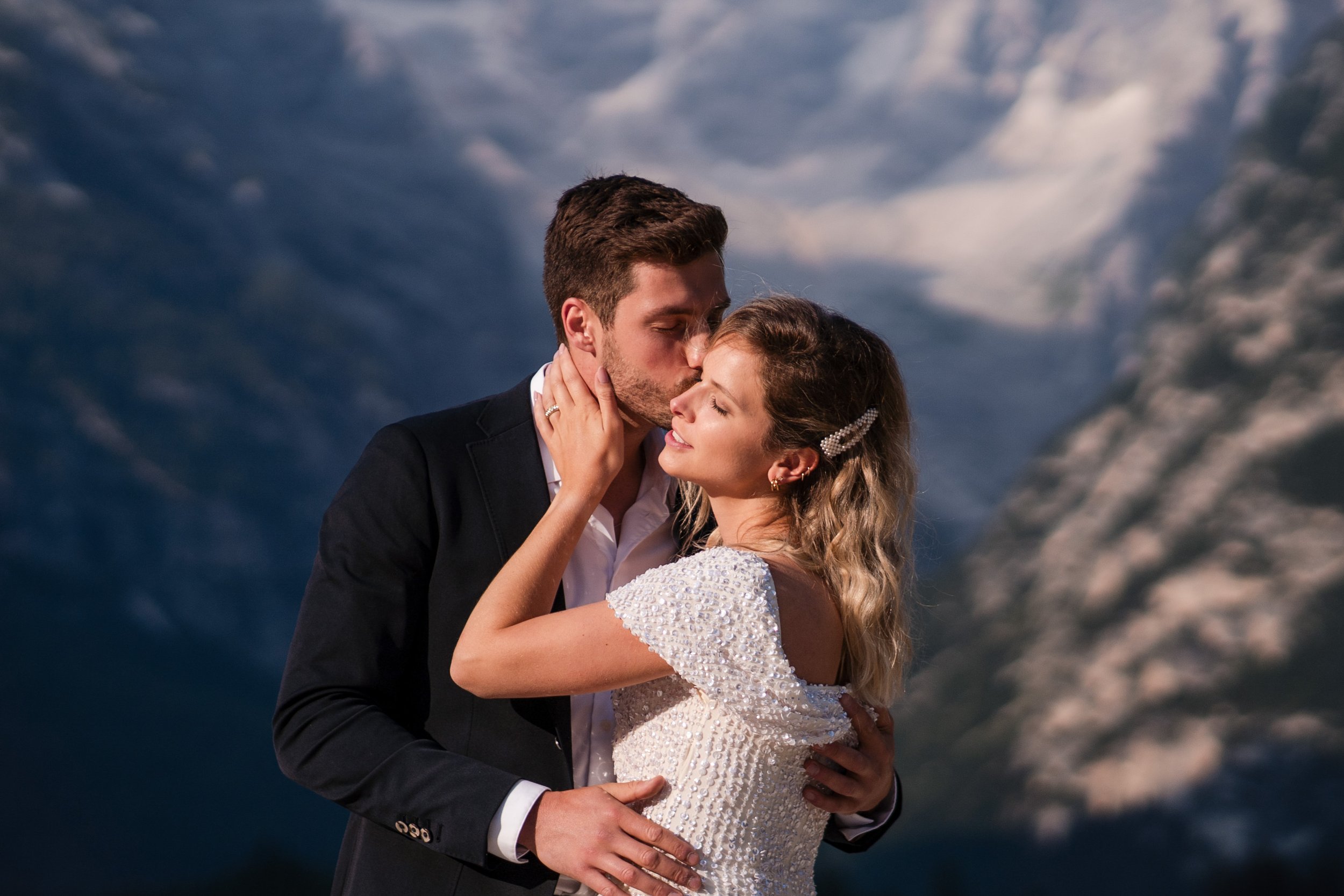 Ein Paar bei einer romantischen Umarmung und Kuss vor einer erdrückenden Bergkulisse, vermutlich während einer Hochzeitsfeier.