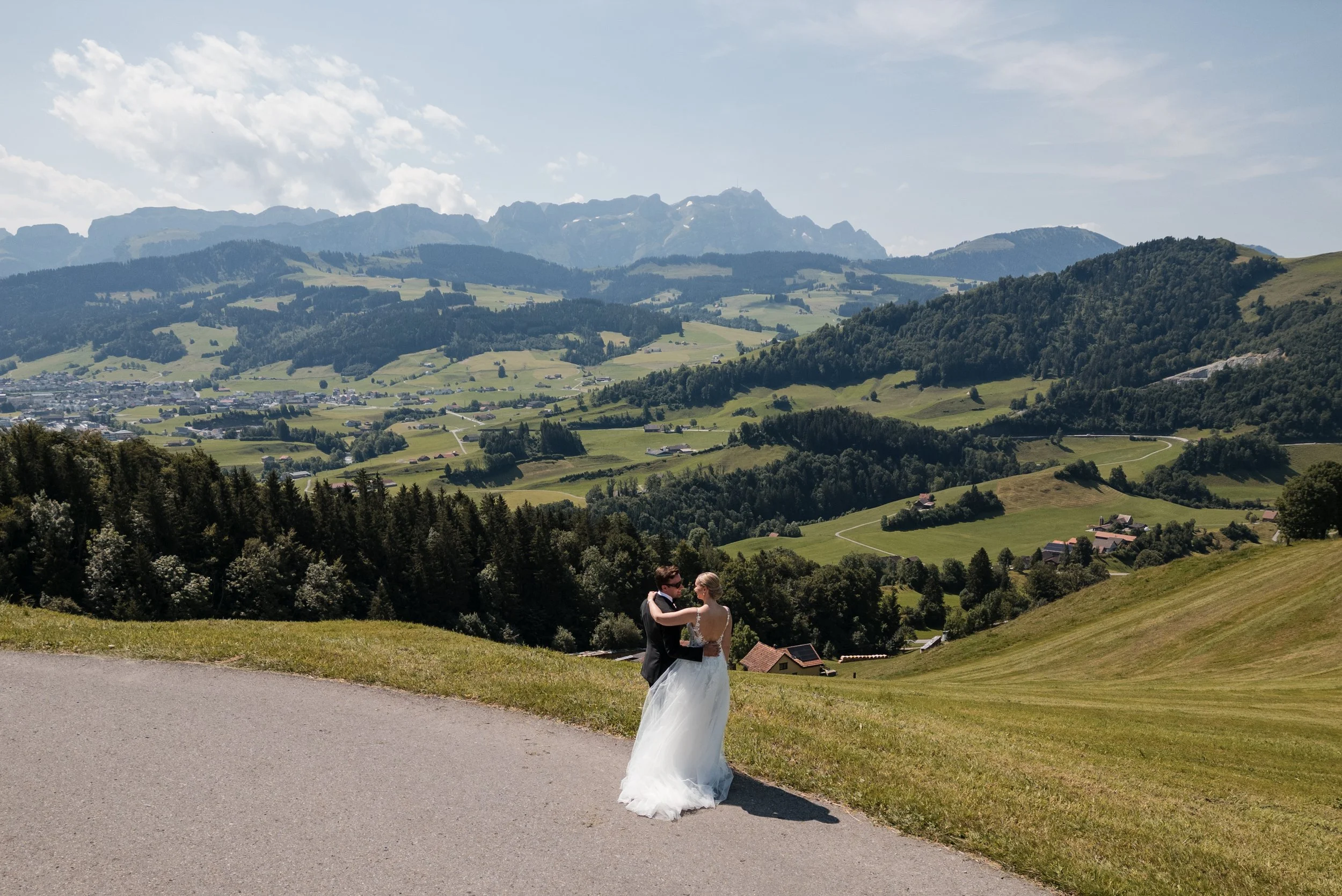 Ein Brautpaar tanzt in einer ländlichen Landschaft mit grünen Hügeln und Bergen im Hintergrund an einem sonnigen Tag.