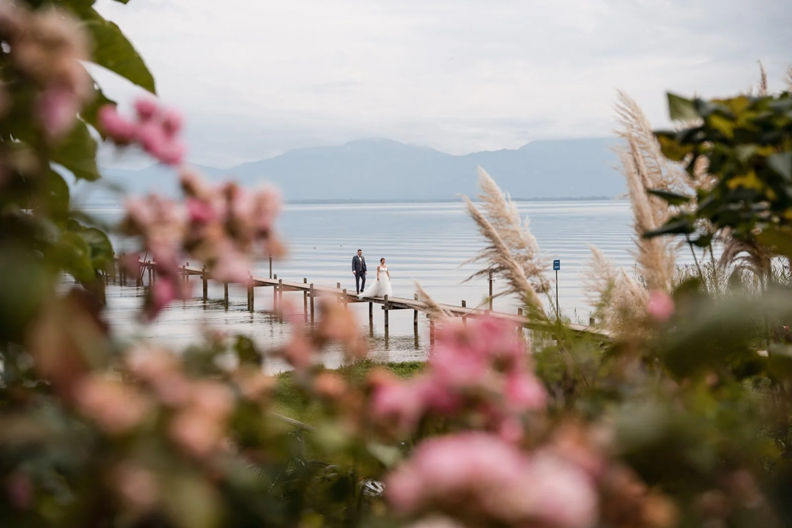 Ein Hochzeitspaar spazier am Steg an einem See, umgeben von Blumen und Gräsern, mit Bergen im Hintergrund.