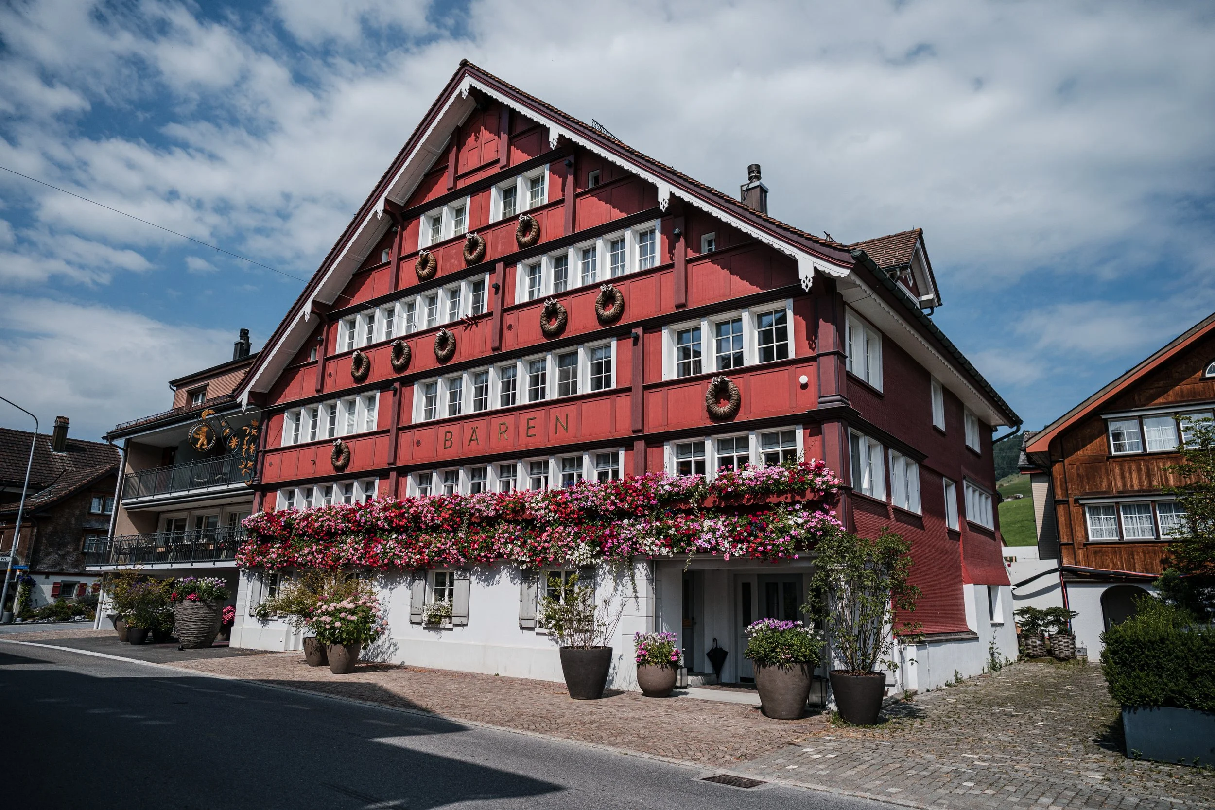 Ein mehrstöckiges, rotes Fachwerkhaus mit weißer Basis, blühenden Blumen und Pflanzkübeln an der Fassade, unter blauem Himmel mit Wolken.