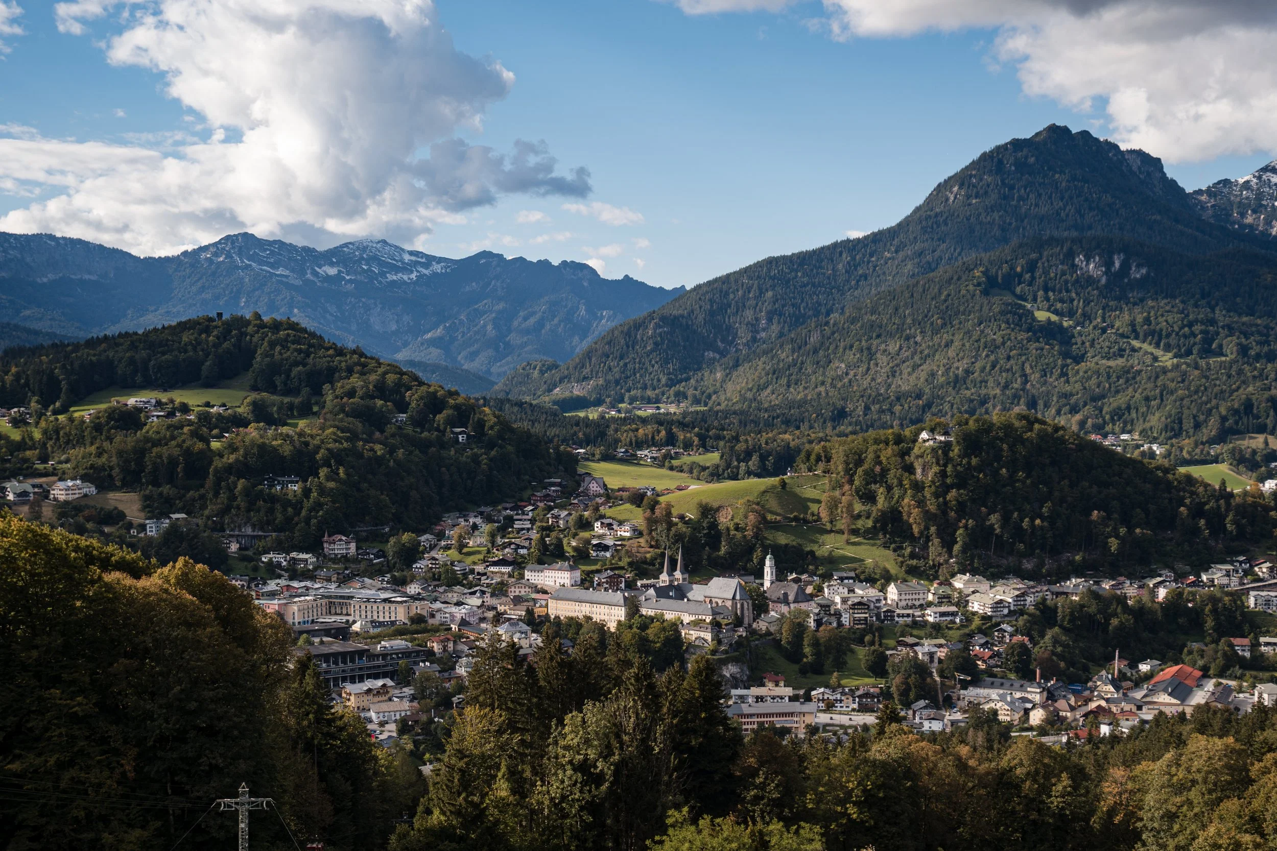 Blick auf eine Stadt im Tal, umgeben von grünen Hügeln und hohen Bergen mit Wolken am Himmel.