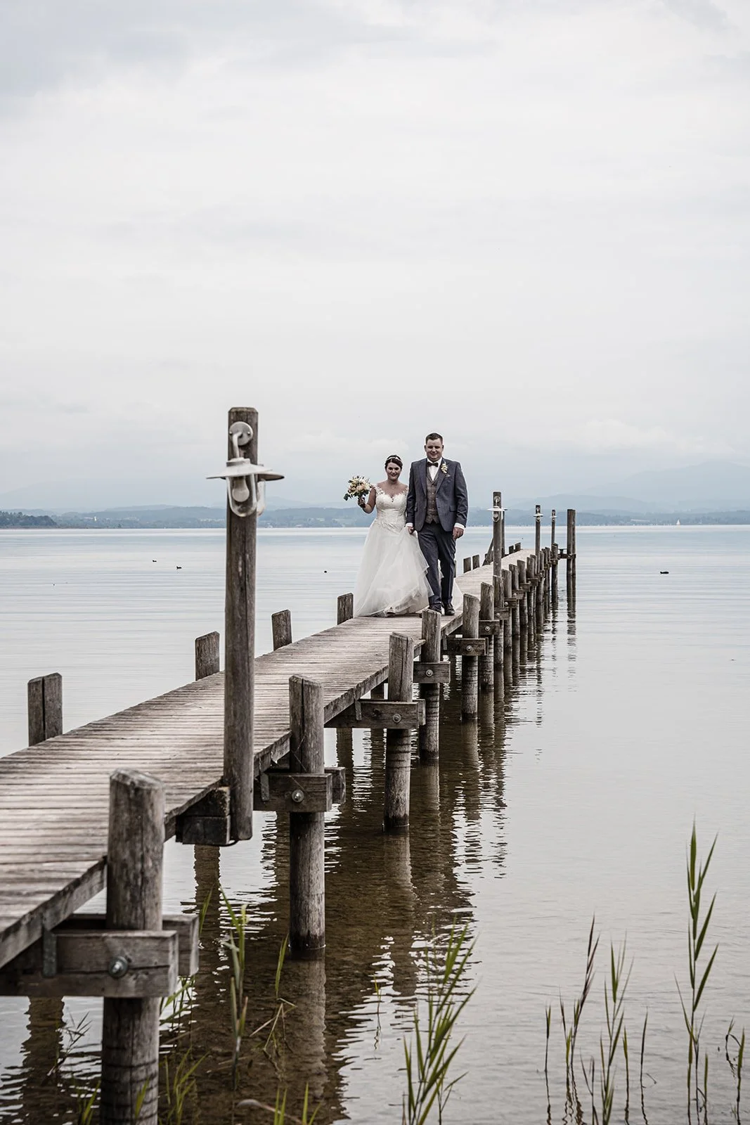 Ein Brautpaar spaziert auf einem hölzernen Steg über einen See, umgeben von Wasser und einer bewölkten Himmel