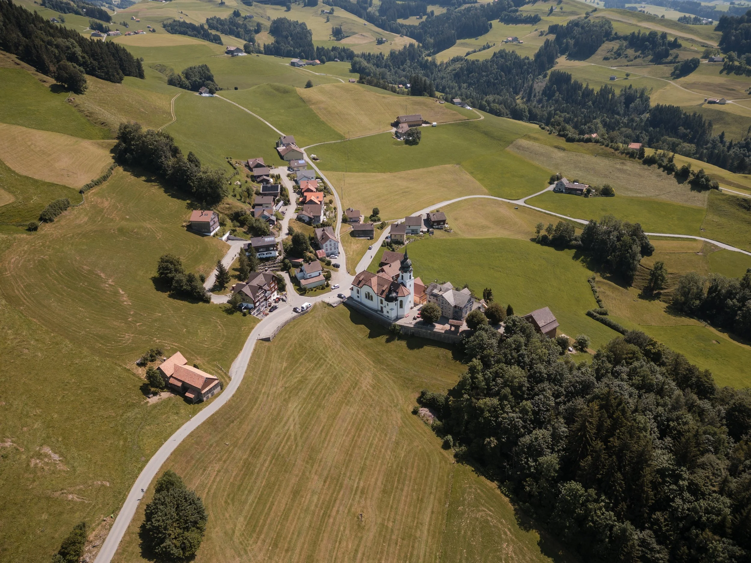 Luftaufnahme eines kleinen Dorfes in einer ländlichen, grünen Landschaft mit Hügeln, Wäldern und einer Kirche im Zentrum.