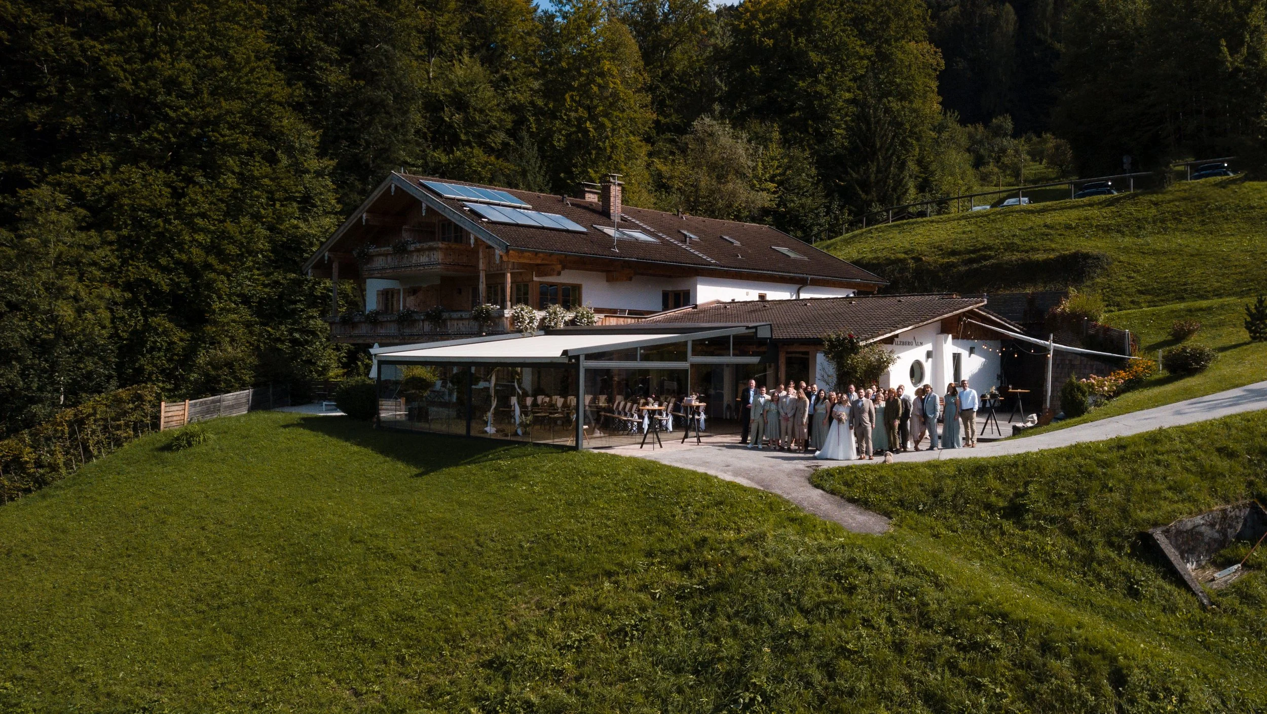 Ein Haus in den Bergen mit einem großen Familien- oder Hochzeitsfest im Freien, mit vielen Menschen, die zusammenstehen, unter einem weißen Pavillon, umgeben von grünem Rasen und Bäumen im Hintergrund.
