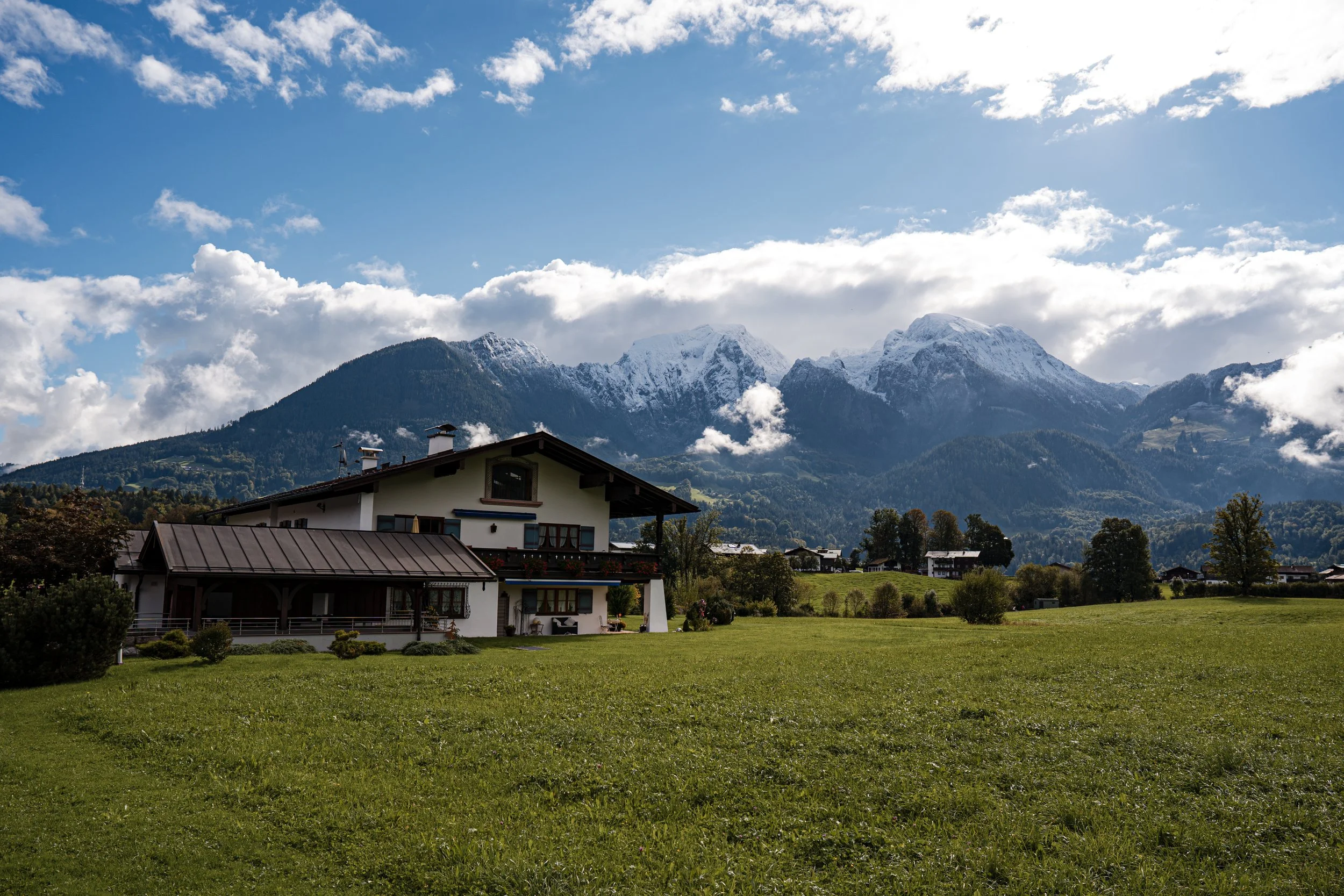 Eine grüne Wiese mit einem Haus im Vordergrund, im Hintergrund schneebedeckte Berge, unter einem blauen Himmel mit Wolken.