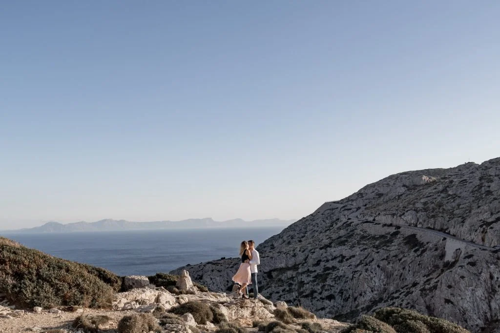 Ein Paar steht auf einer felsigen Küstenlandschaft mit Blick auf das Meer und Berge im Hintergrund.