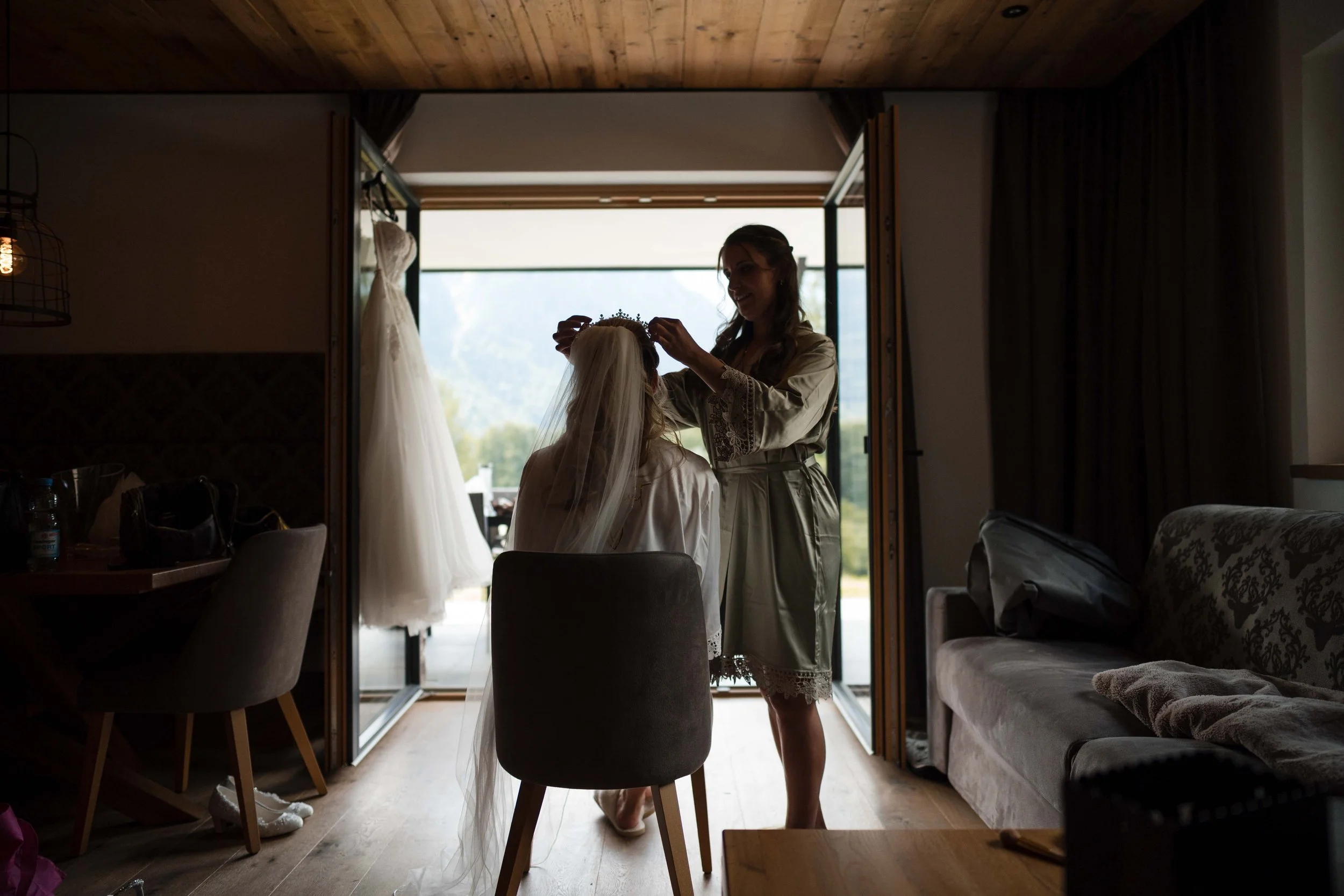 Eine Braut wird von einer Frau in einem Raum vor einem offenen Fenster mit Blick auf eine ländliche Landschaft frisiert, wobei die Braut ein Brautkleid und Schleier trägt.