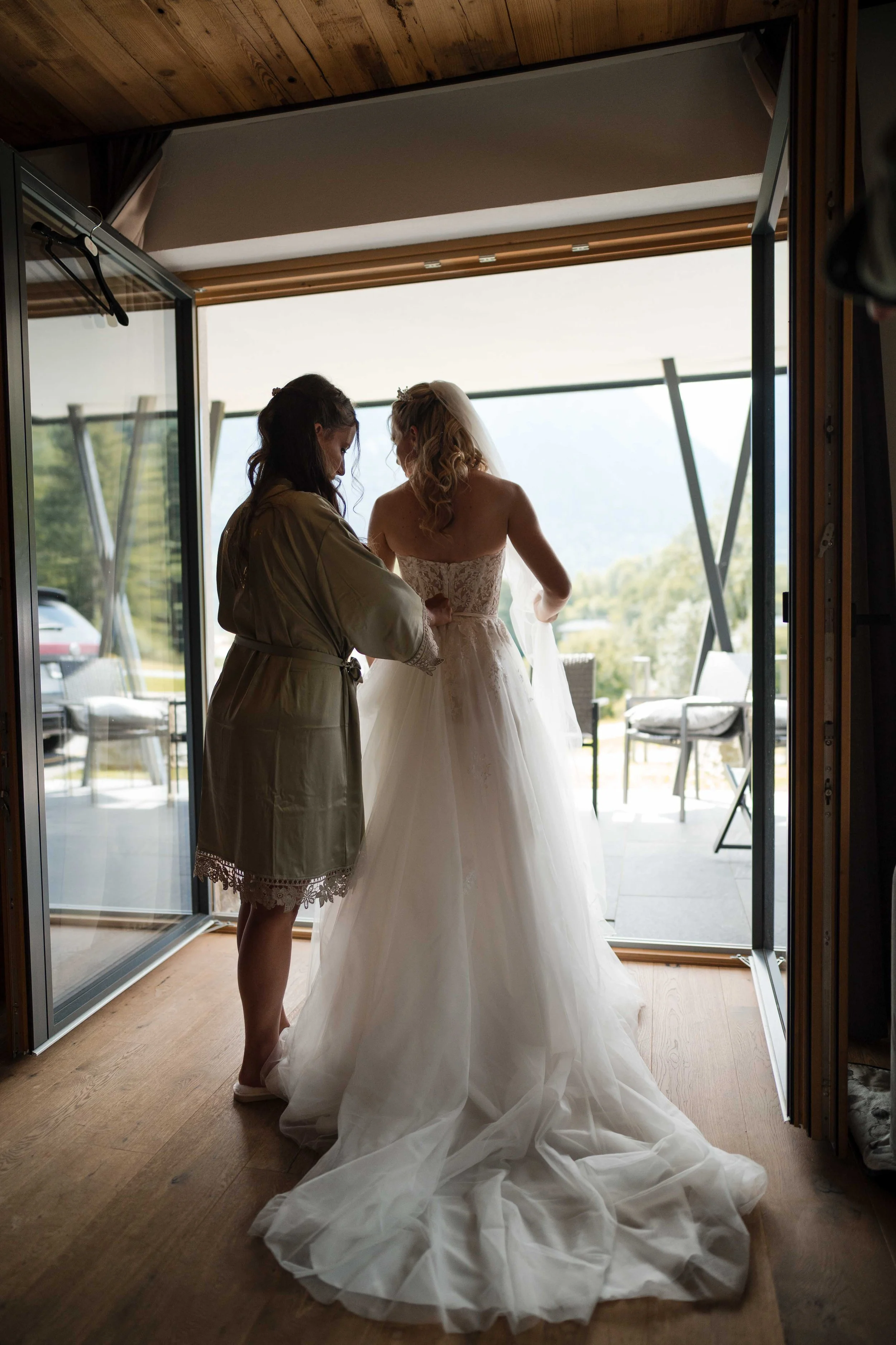 Braut wird von einer Helferin vor der Hochzeit in einem Raum vorbereitet, Blick nach draußen auf einen Balkon mit Stühlen und Blick in die Landschaft.