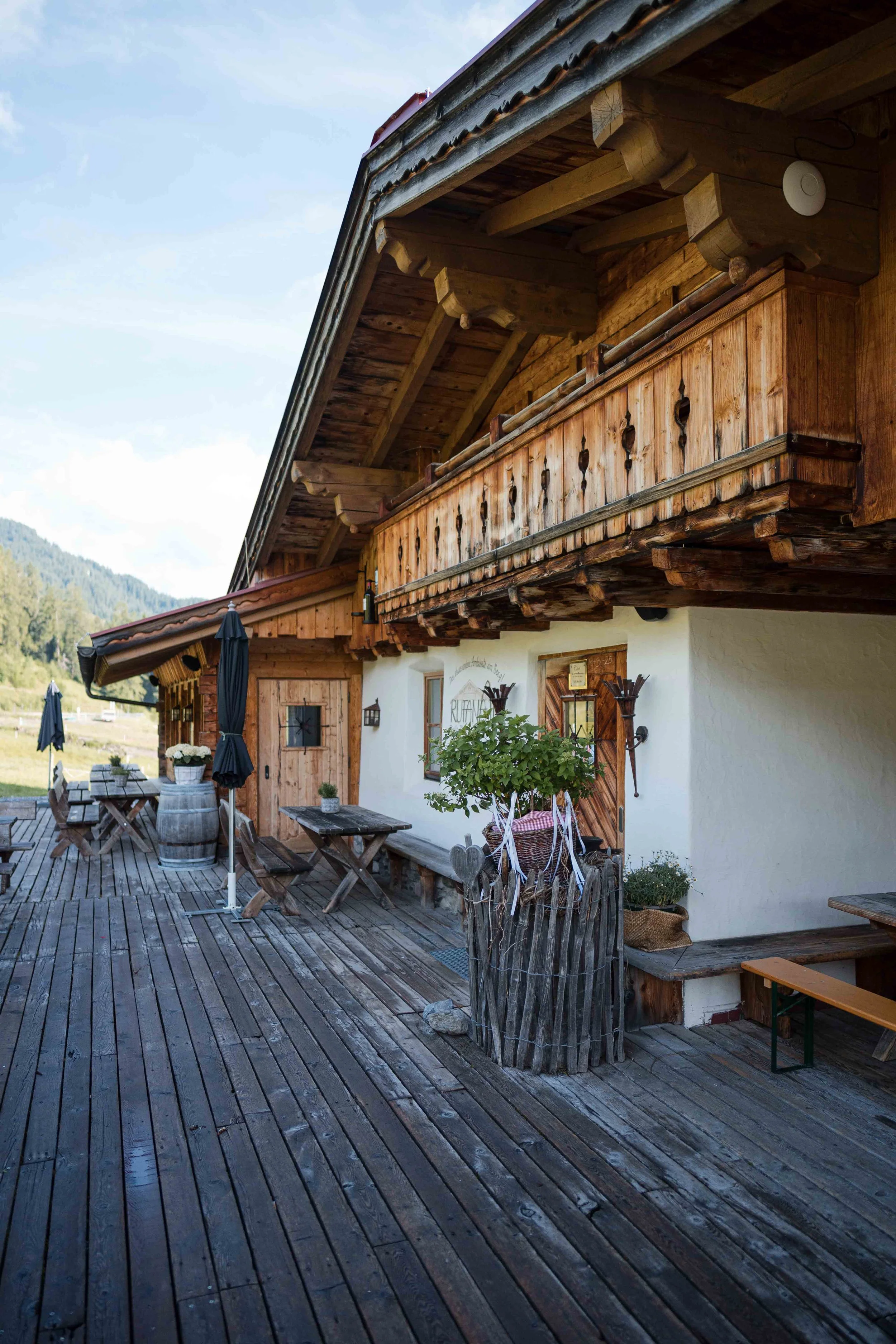 Blick auf eine traditionelle Holzhütte mit Balkon und Terrasse im alpinen Stil, mit Sitzgelegenheiten und Sonnenschirmen auf einer Holzterrasse, umgeben von grüner Natur und Bergen im Hintergrund.
