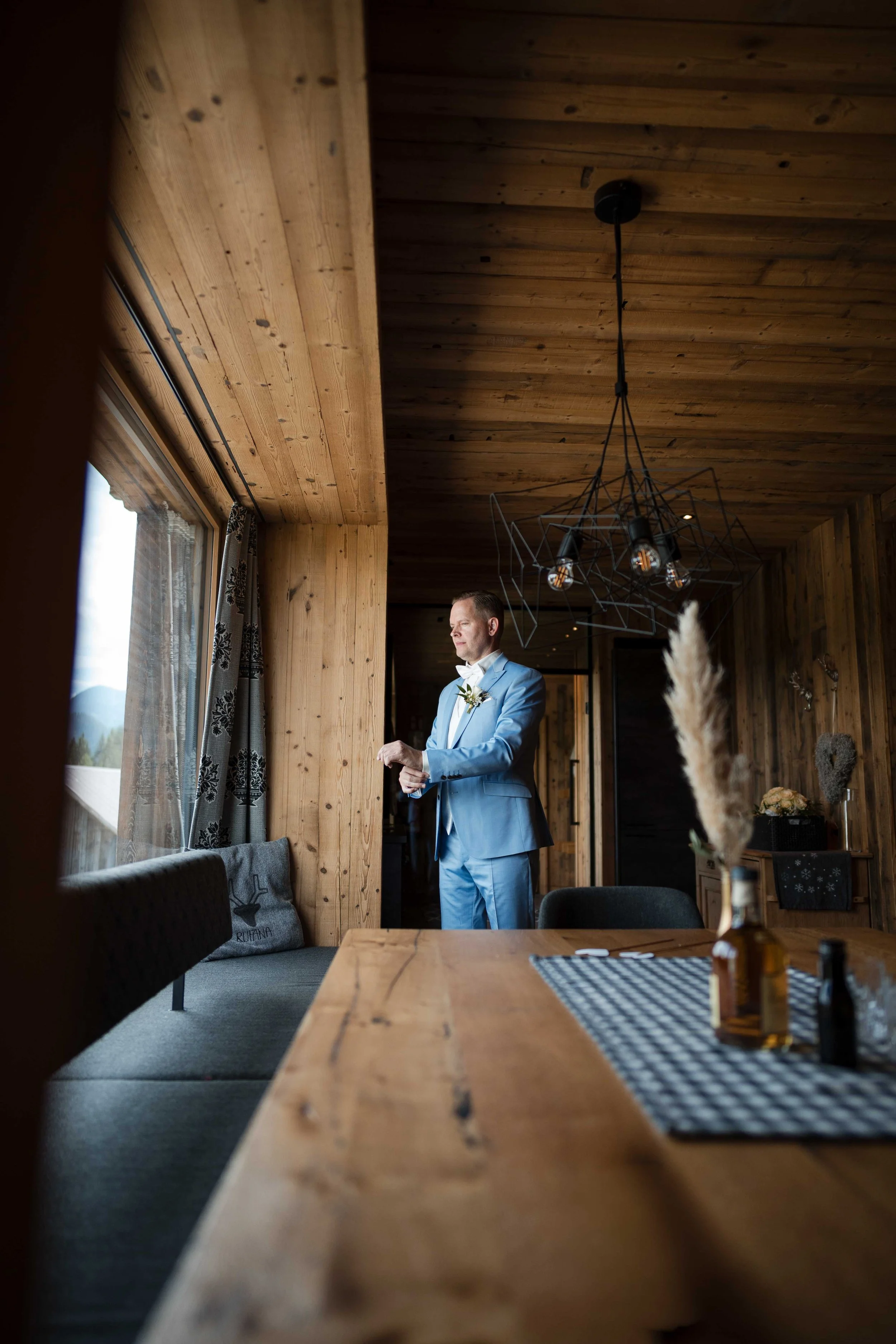 Ein Mann in einem hellblauen Anzug steht im Wohnzimmer eines Holzhauses und schaut aus dem Fenster. Der Raum ist mit Holz verkleidet und hat eine moderne Leuchte an der Decke. Im Vordergrund steht ein Holztisch mit Kerzen und Deko. An der Wand hängt 