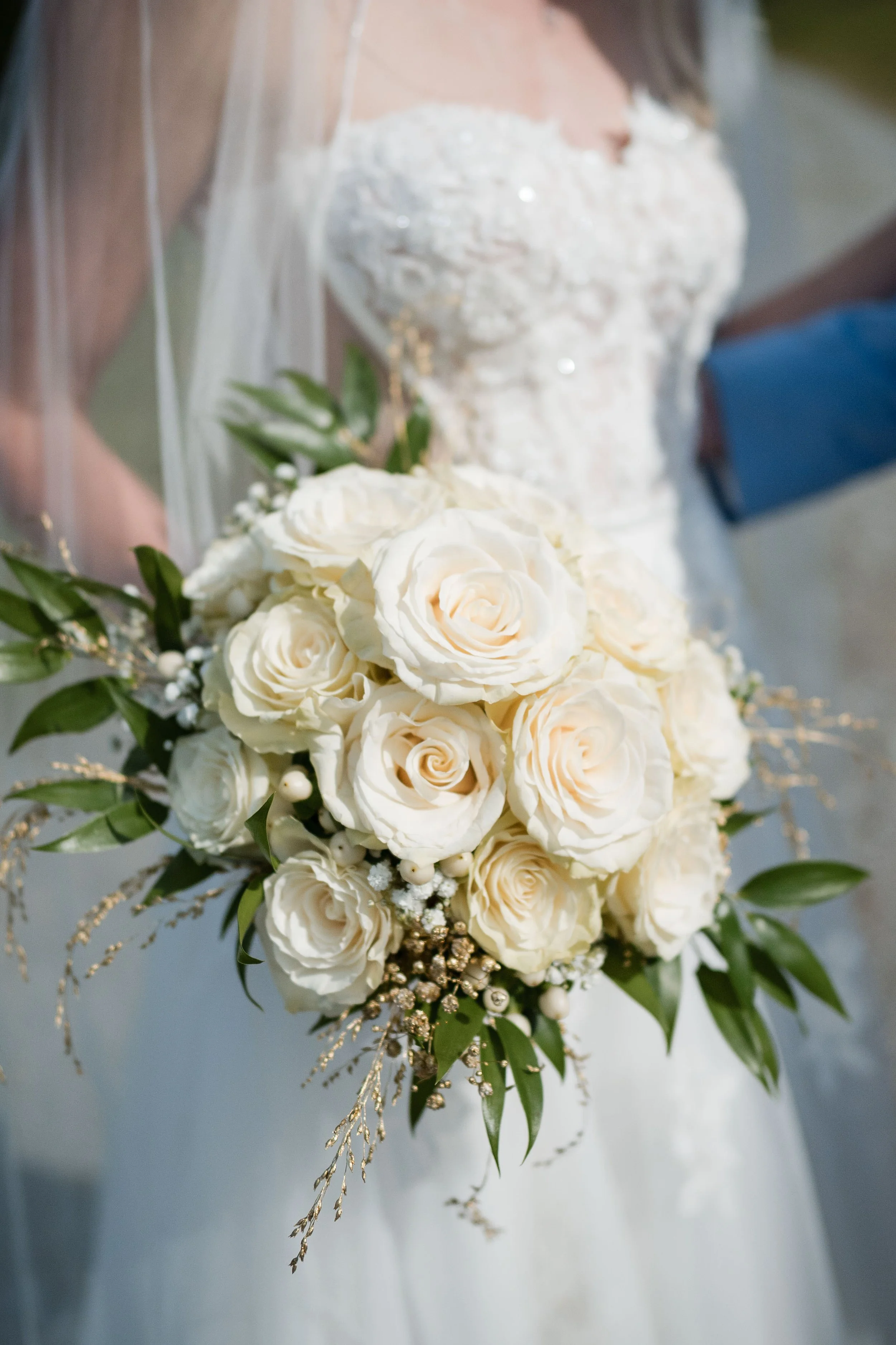 Braut in Hochzeitskleid hält einen weißen Brautstrauß aus Rosen mit grünen Blättern und Dekorationen.