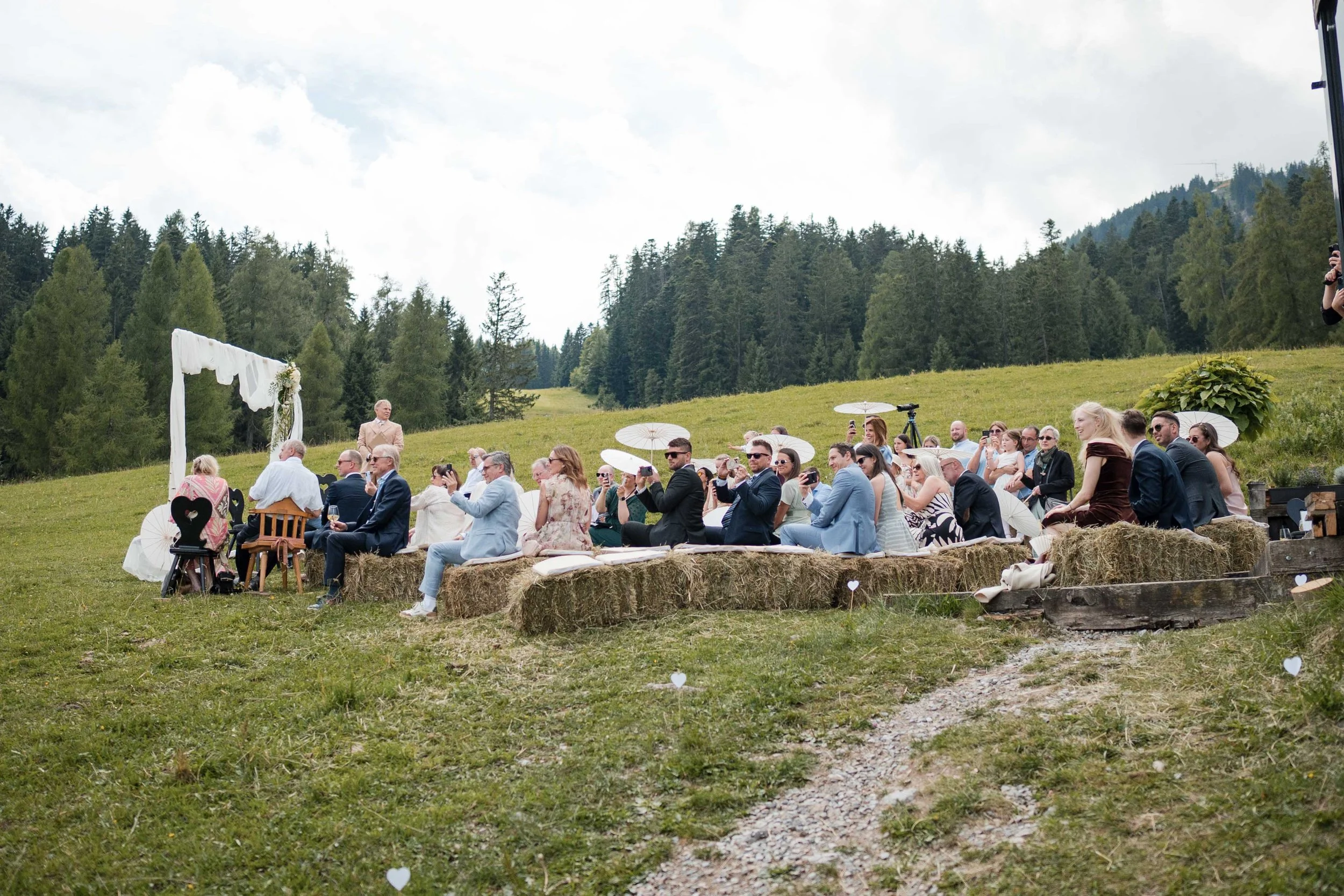 Hochzeit im Freien mit Gästen, die auf Heuballen sitzen, in einer grünen, bergigen Landschaft, einige halten Regenschirme, ein weißer Hochzeitsbogen ist im Hintergrund sichtbar.