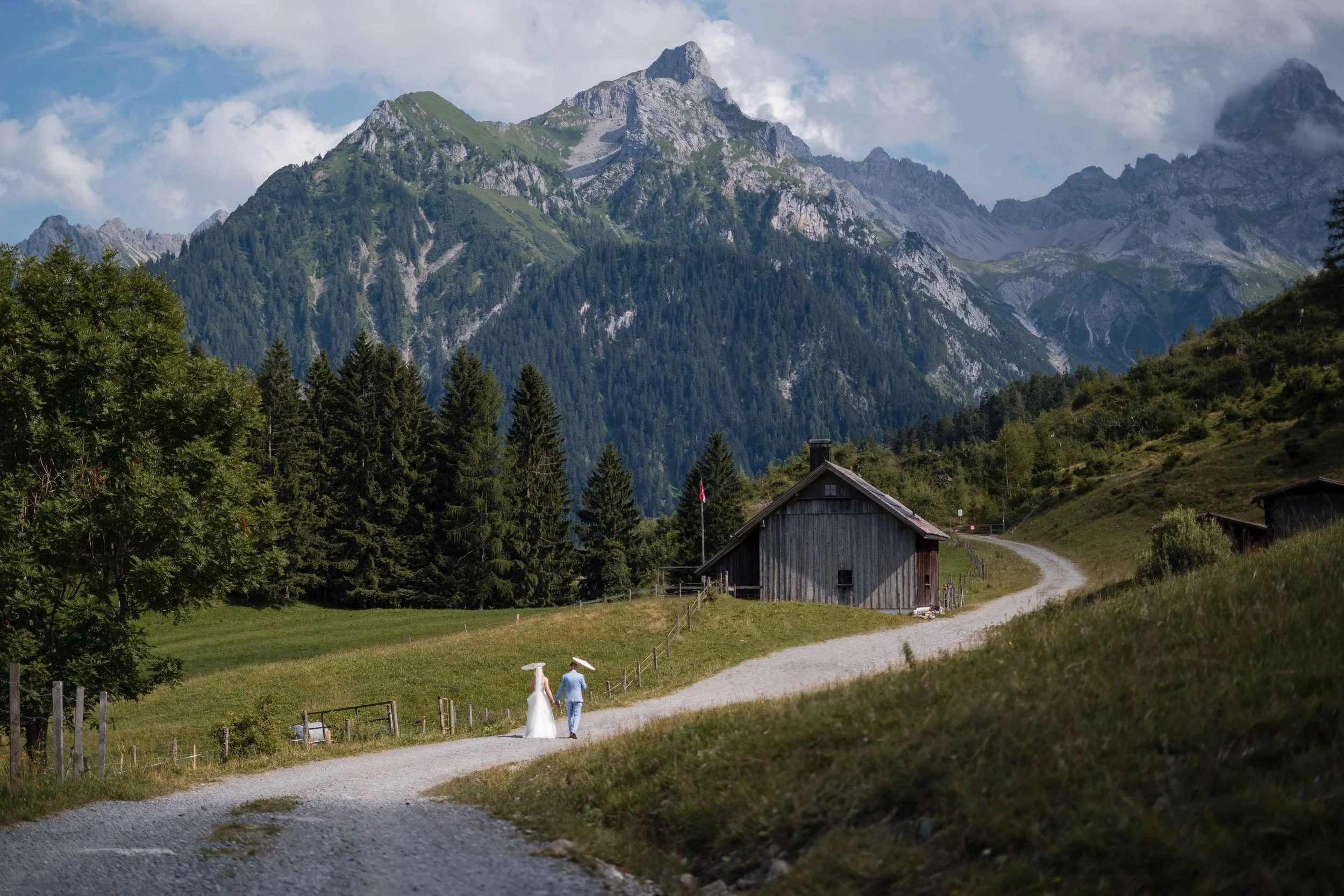 Ein Brautpaar wandert auf einem schmalen Weg in einer ländlichen, bergigen Landschaft, umgeben von Wäldern und Bergen.
