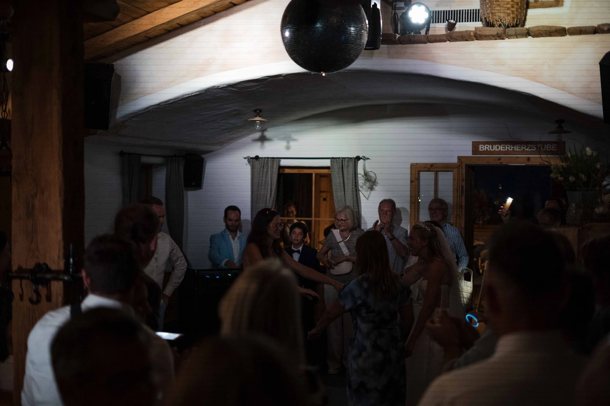 Menschen feiern in einer rustikalen Holzhütte, einige tanzen, andere beobachten, mit einem Disco-Kugel an der Decke, festliche Beleuchtung und Blumen im Hintergrund.