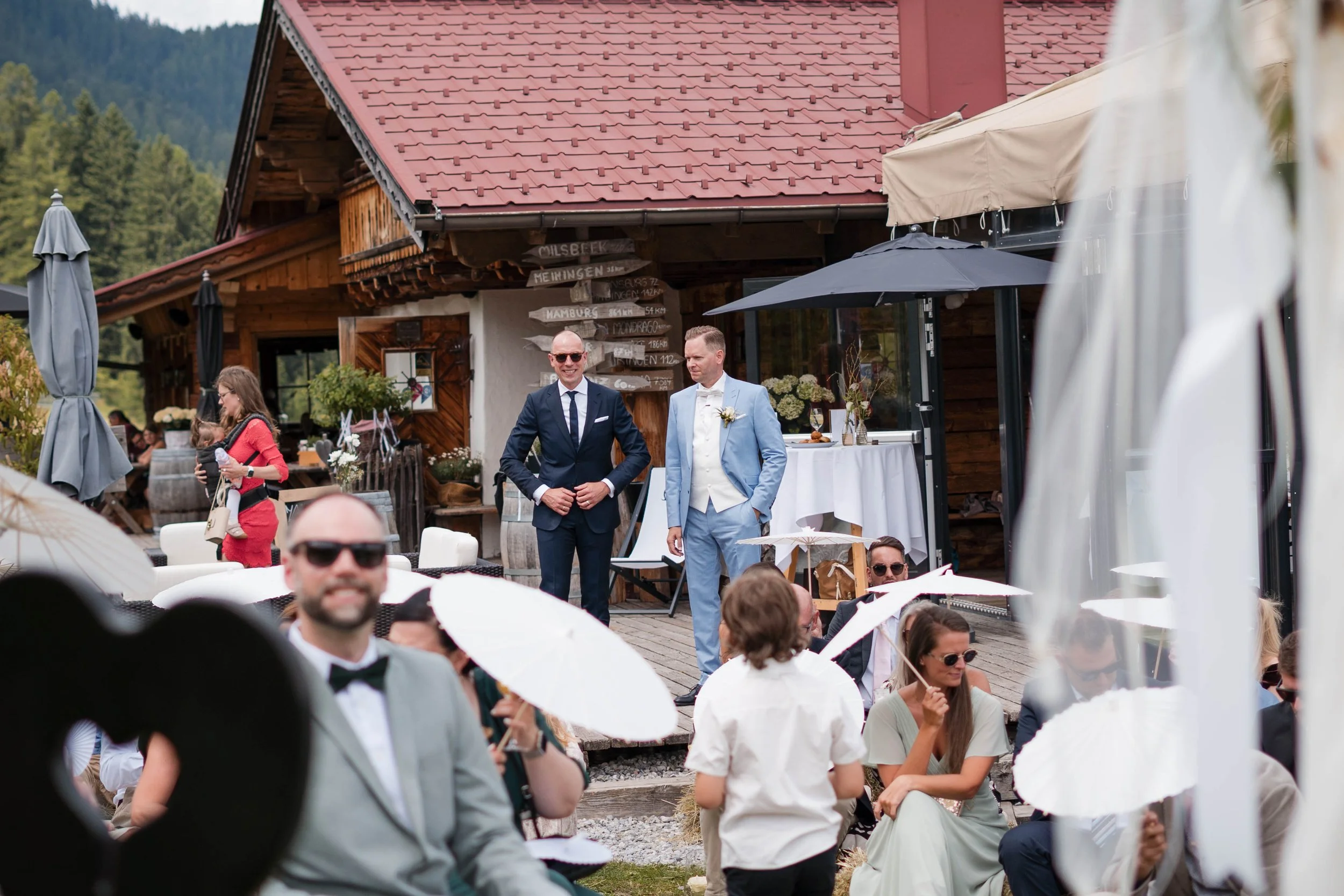Viele Gäste bei einer Hochzeit im Freien, mit weißen Regenschirmen, sitzen und stehen vor einem Holzhaus, im Hintergrund sind Bäume und Berge, zwei Männer beim Reden, eine Frau mit einem Kind, alle in formeller Kleidung.