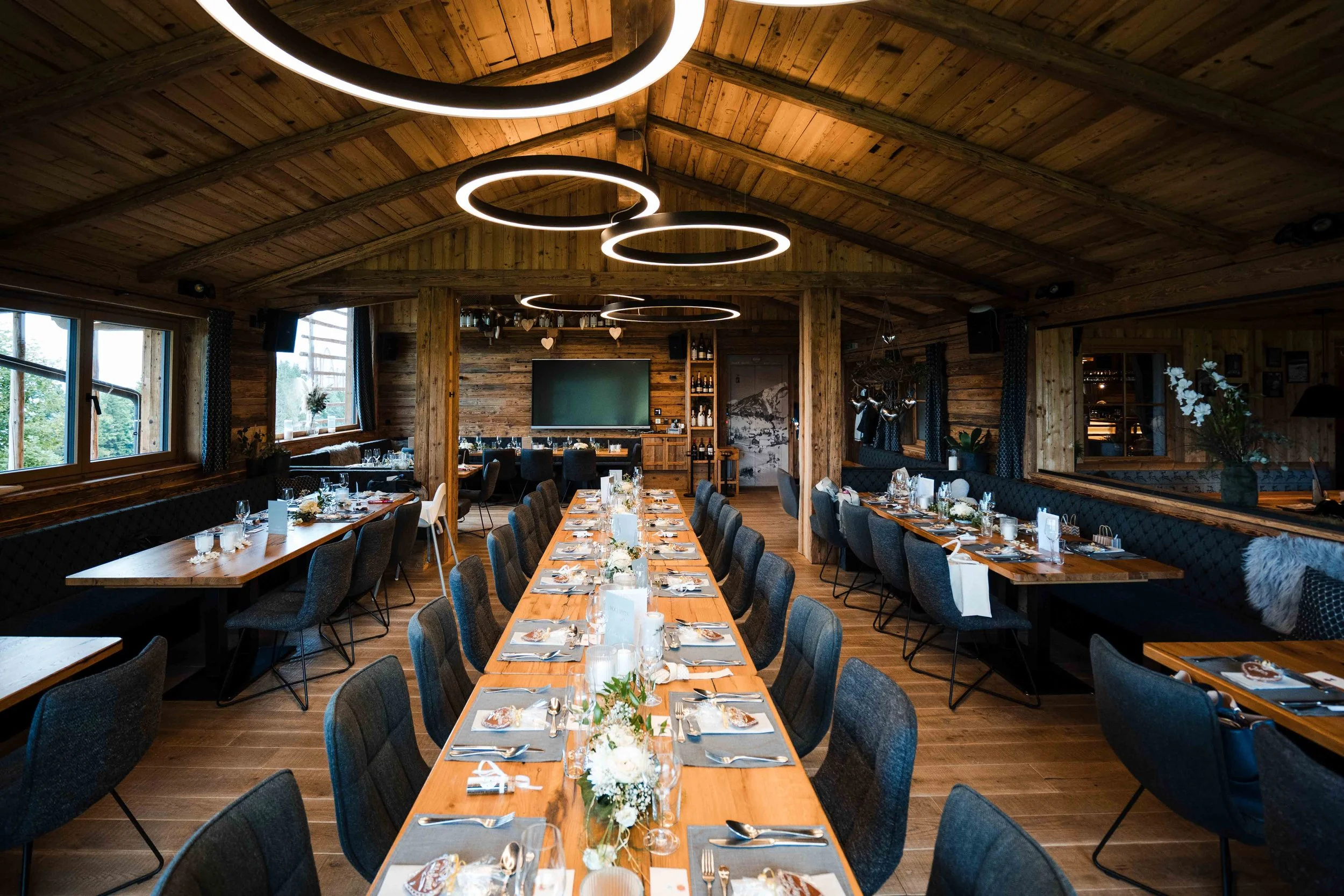 Ein festlich gedeckter Raum mit langen Holztischen, Kerzen und Blumen für eine Veranstaltung in einem rustikalen Holzhaus mit moderner Beleuchtung.