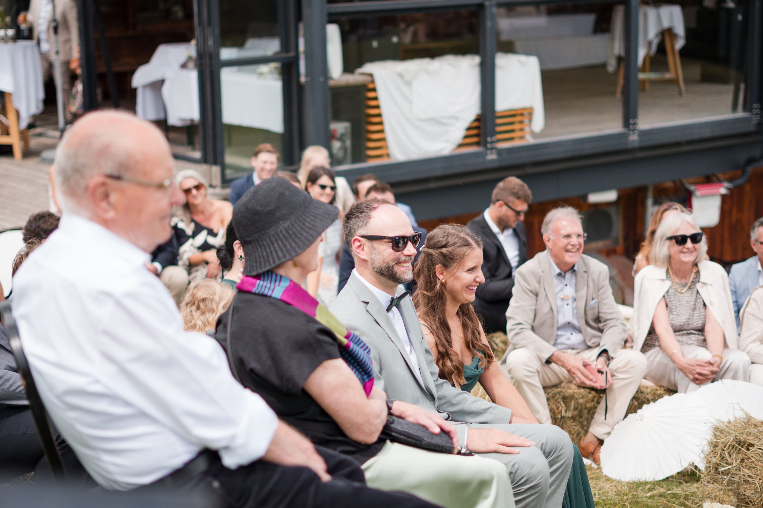Menschen bei einer outdoor Veranstaltung, wahrscheinlich eine Hochzeit, sitzend auf Hay Bales, einige tragen Sonnenbrillen, lächelnd, im Hintergrund ein modernes Gebäude.