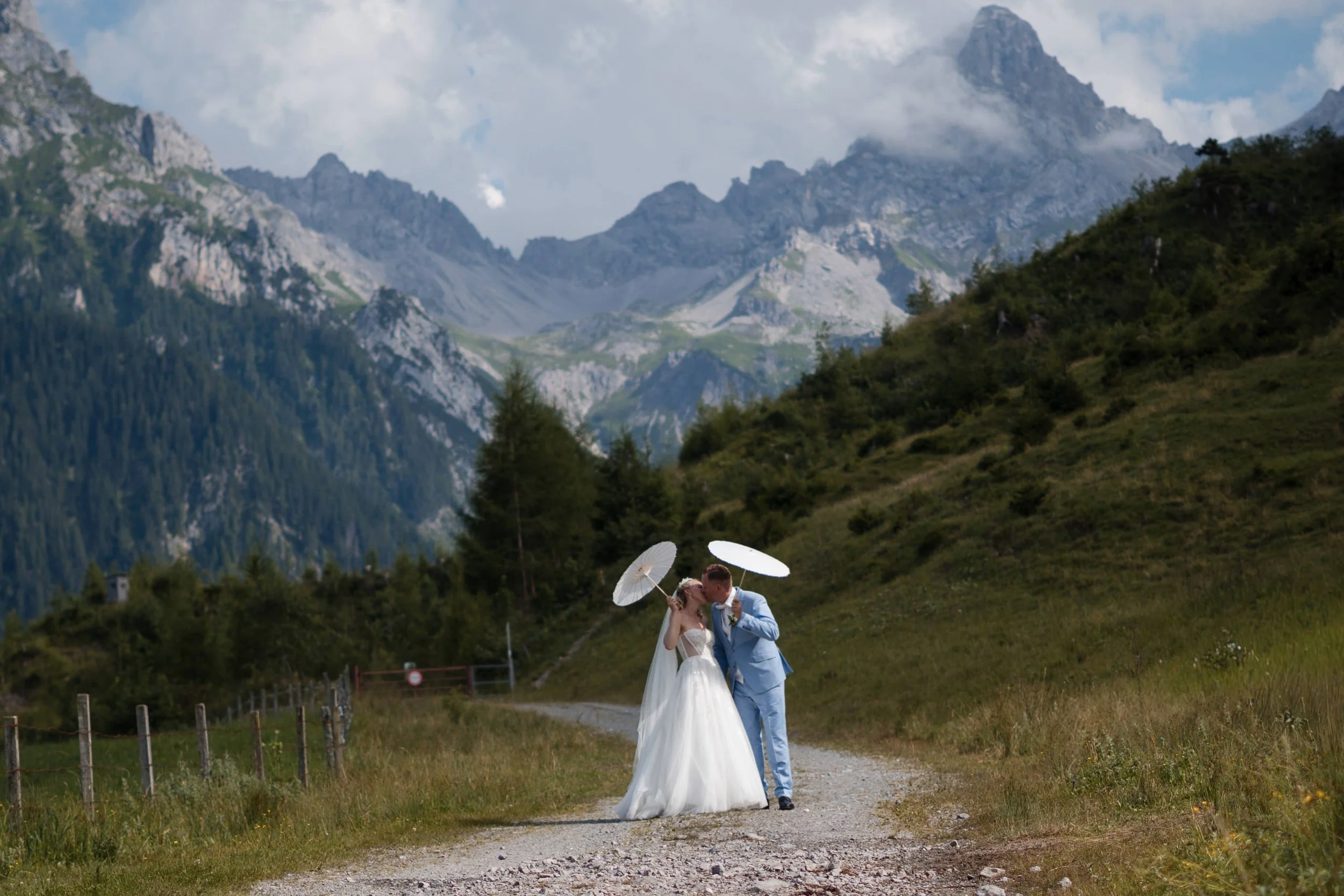 Ein Brautpaar in Hochzeitskleidung, der Braut in einem weißen Kleid und der Bräutigam in einem blauen Anzug, stehen auf einem Weg in einer bergigen Landschaft, umgeben von grünen Wiesen und hohen Bergen im Hintergrund. Sie halten weiße Handregen und 