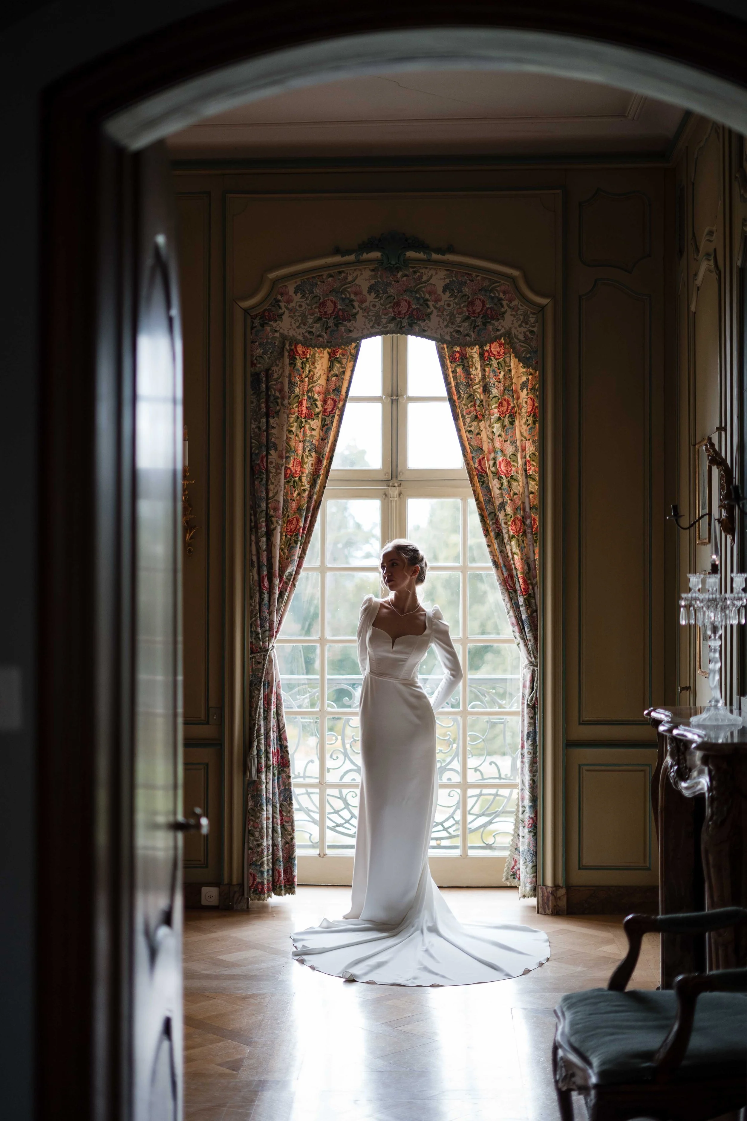 Braut in einem weißen Kleid steht vor einem großen Fenster mit blumenverzierten Vorhängen in einem eleganten, históricoen Raum.