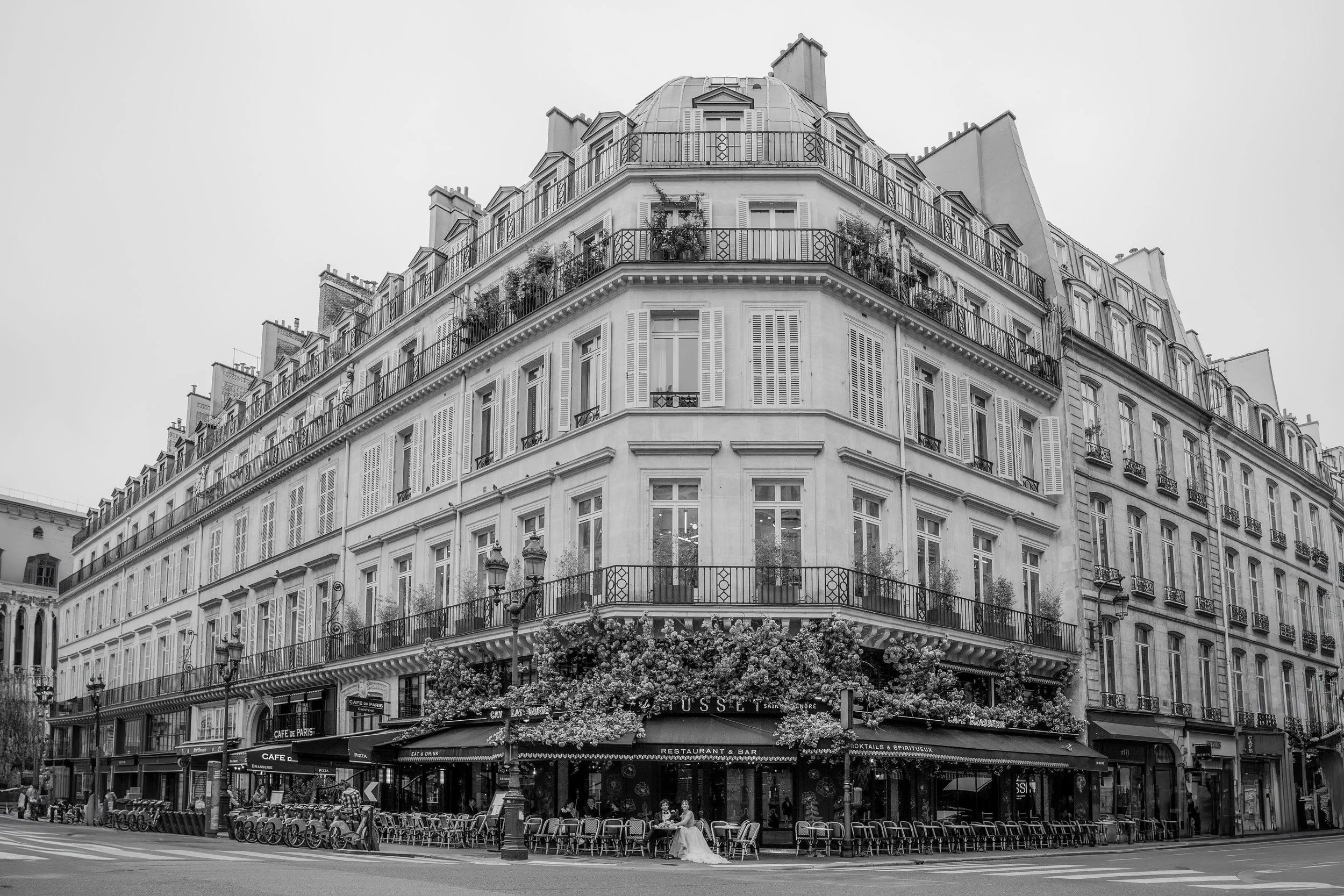 Ein schwarzes und weißes Foto eines mehrstöckigen Stadthauses mit Cafés und Restaurants im Erdgeschoss, zahlreiche Fenster mit Balkon- und Blumendekorationen, Menschen auf der Straße und an Tischen