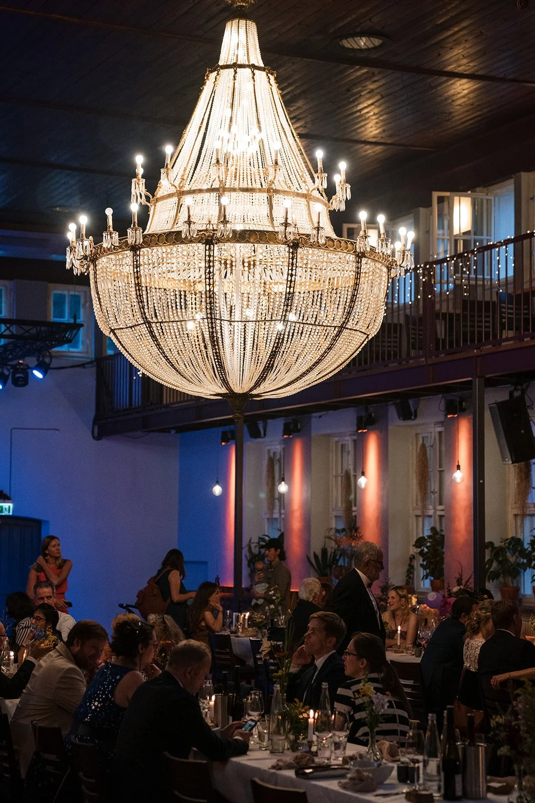 Ein eleganter Kronleuchter hängt in einem großen Raum während einer formellen Veranstaltung. Im Hintergrund sind Gäste an Tischen, die sich unterhalten und speisen, sichtbar.