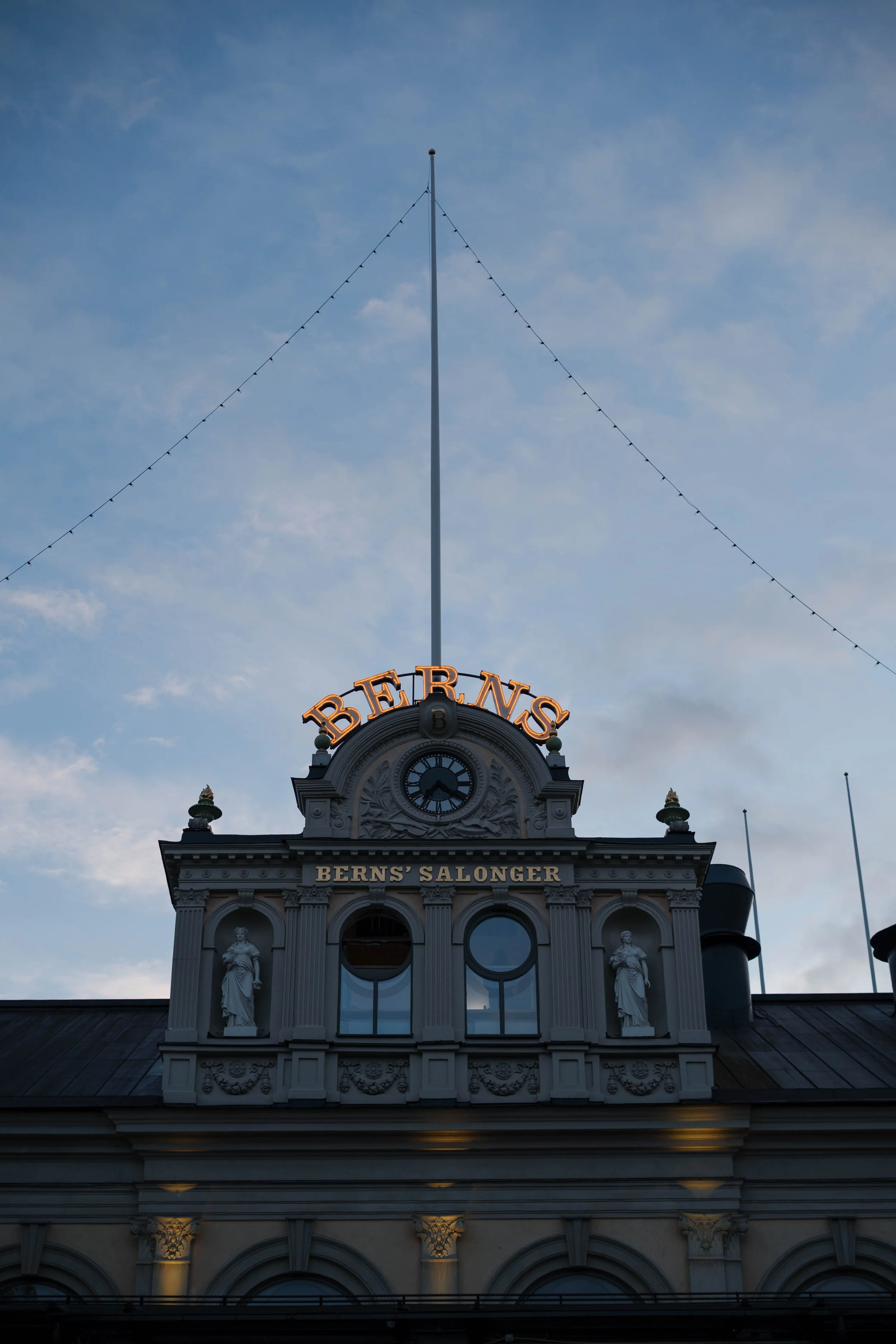 Ansicht eines historischen Gebäudes mit dem Schriftzug 'BERNS' und einer Uhr, bei Dämmerlicht, mit Lichtern am Gebäude und himmelblauem Himmel im Hintergrund.