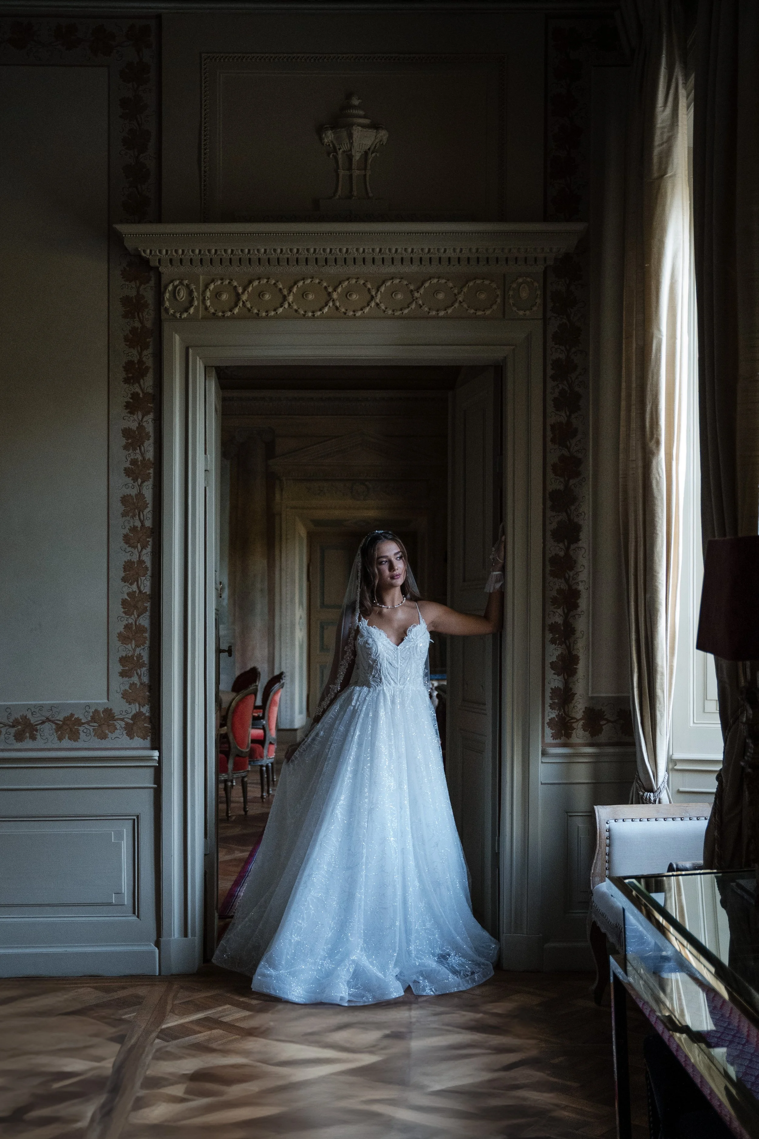 Braut in einem weißen Hochzeitskleid steht in einem eleganten, historischen Raum mit großen Fenstern und schweren Vorhängen.