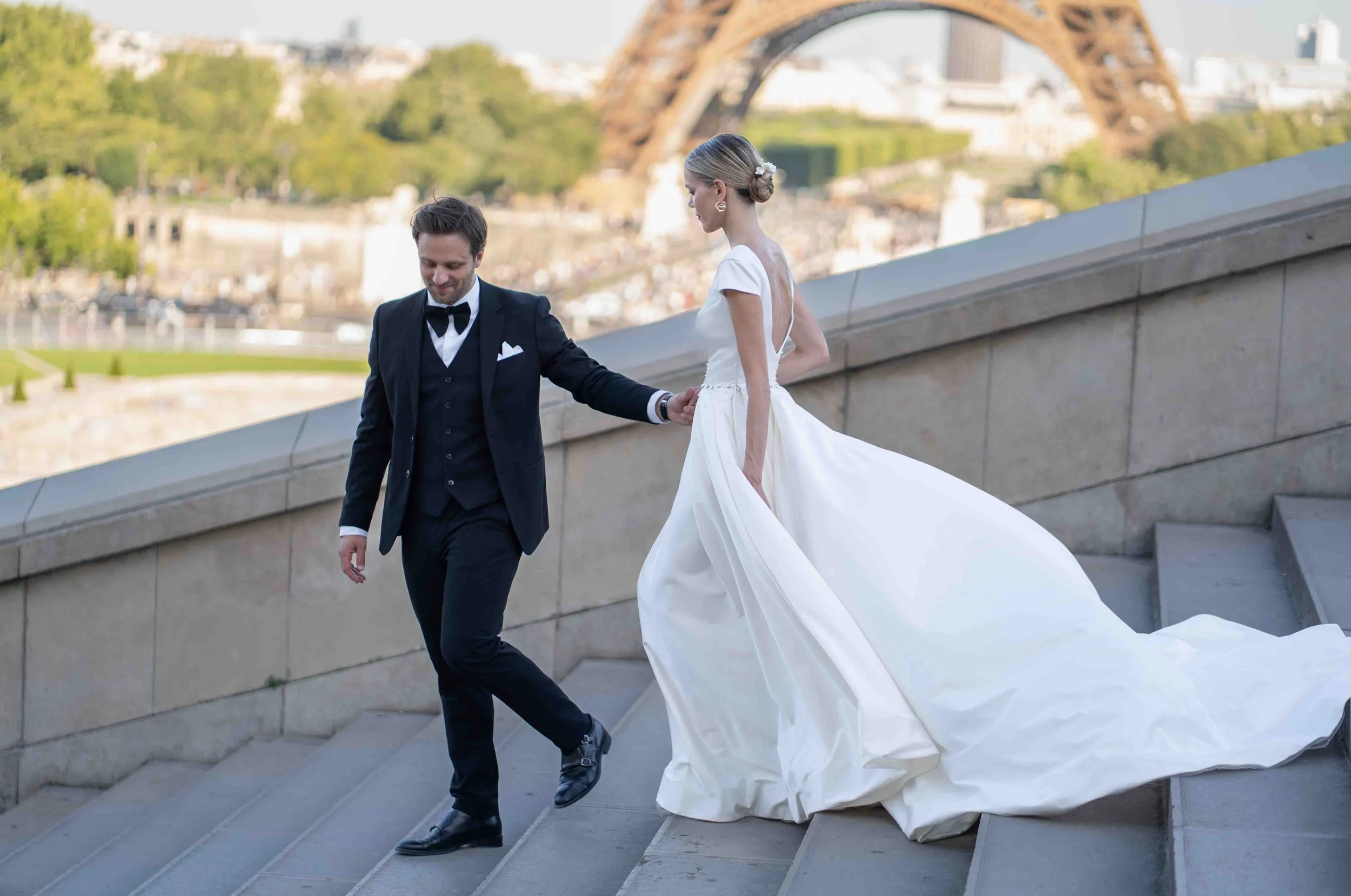 Ein Brautpaar auf Treppen in Paris mit Blick auf die Stadt, die Eiffel-Turm im Hintergrund, bei ihrer Hochzeitszeremonie. Der Bräutigam trägt einen schwarzen Anzug mit Fliege, die Braut in einem weißen Hochzeitskleid.