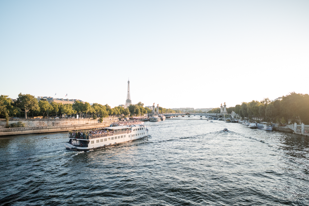 Blick auf die Seine mit einem Kreuzfahrtschiff, mehreren kleineren Booten, dem Eiffelturm im Hintergrund und Bäumen an den Ufern bei Sonnenuntergang in Paris.