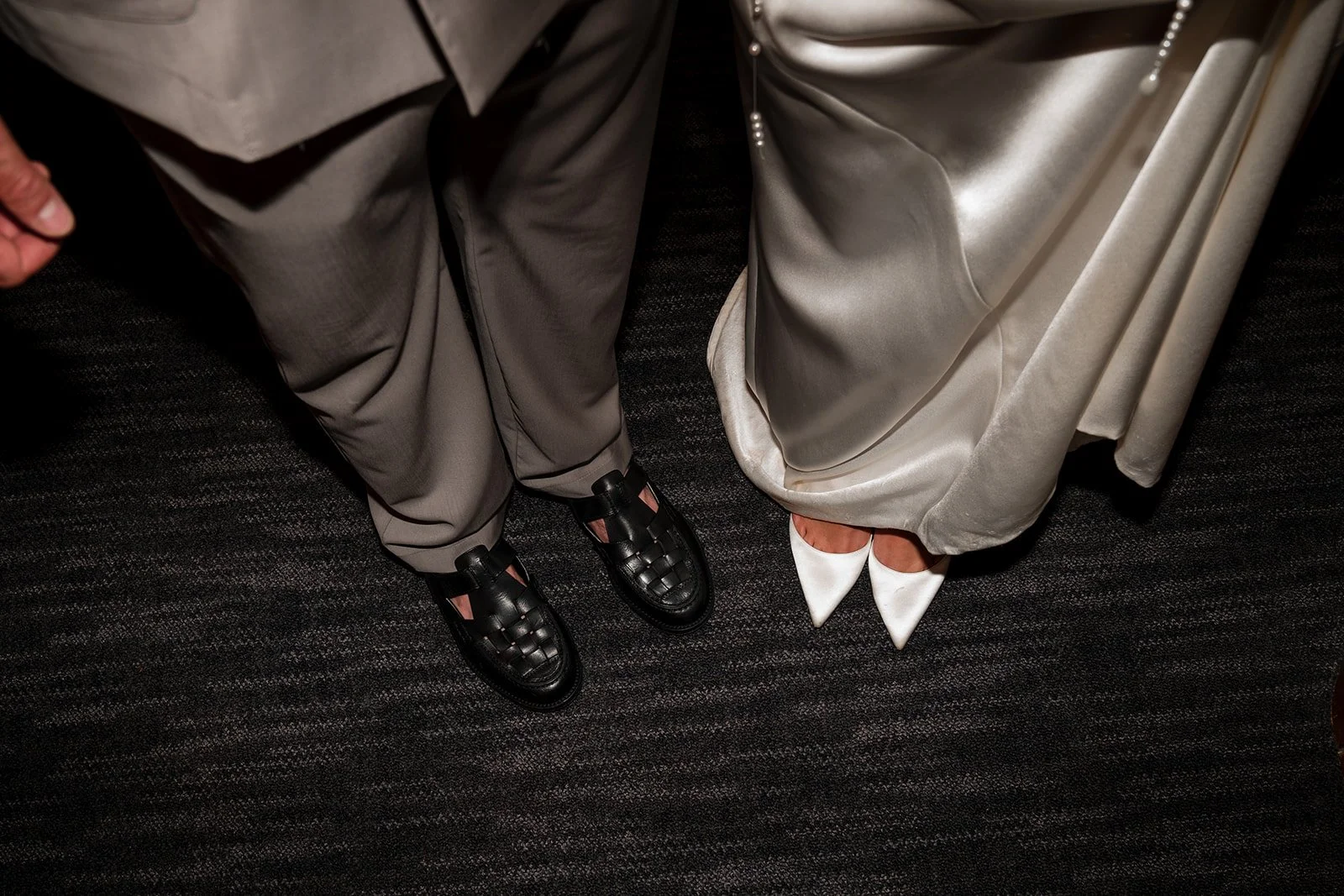 Blick nach unten auf die Füße eines Paares, das formelle Kleidung trägt: die Person auf der linken Seite trägt schwarz-rosafarbene Schuhe und graue Hosen, die Person auf der rechten Seite trägt weiße High Heels und ein langes, cremefarbenes Kleid.