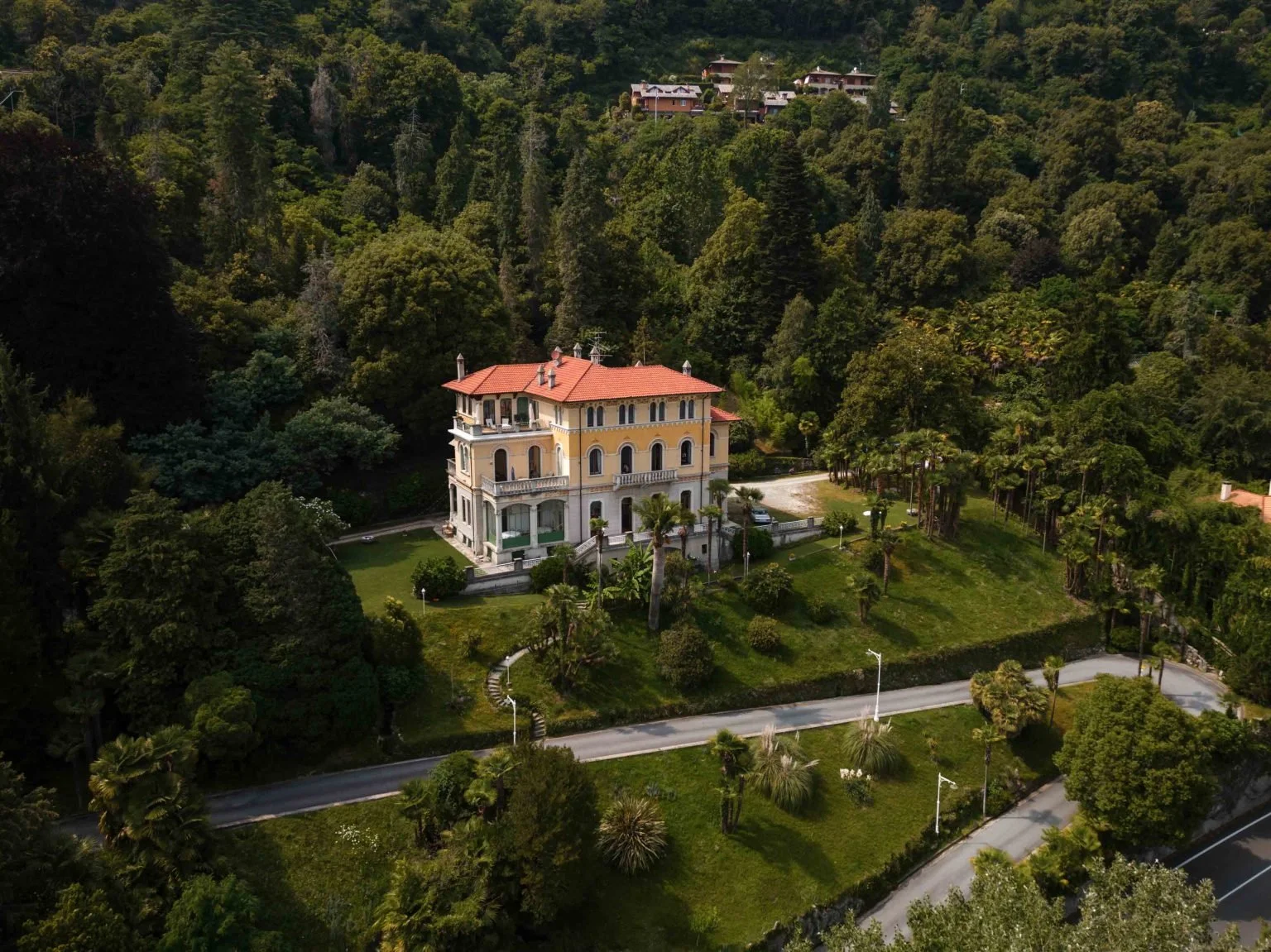 Ein großes, elegantes, mehrstöckiges Haus in einer bewaldeten Gegend, umgeben von gepflegtem Garten und Gehwegen, mit einem norwegischen Stil haus im Hintergrund auf einem Hügel