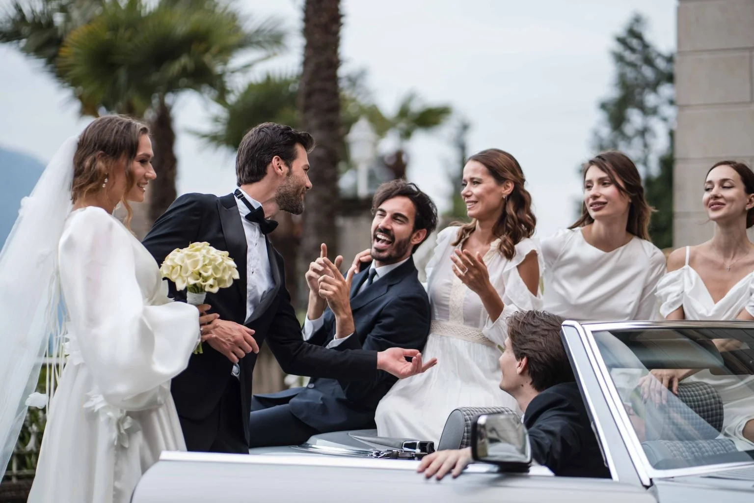 Hochzeitspaar und Gäste feiern in einem weißen Cabrio, das Brautpaar in Brautkleid und Anzug, Gäste in weißen Kleidern, im Freien unter Palmen, fröhliche Stimmung.