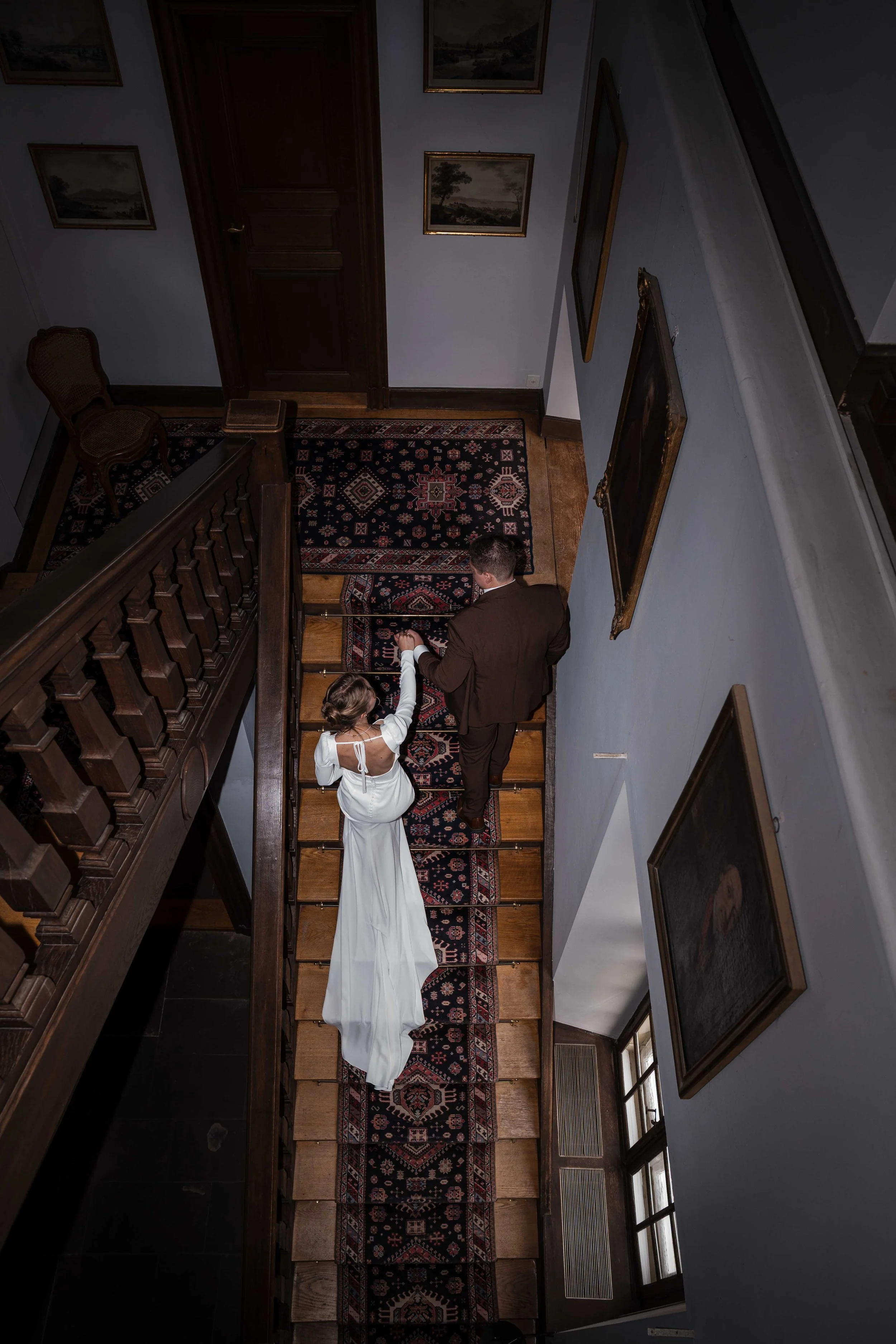 Ein Brautpaar auf einer Treppe in einem alten, gut ausgestatteten Haus. Der Braut trägt ein weißes Hochzeitskleid, der Bräutigam einen dunklen Anzug. Die Treppe ist mit einem Teppich bedeckt, die Wände sind mit Gemälden geschmückt.