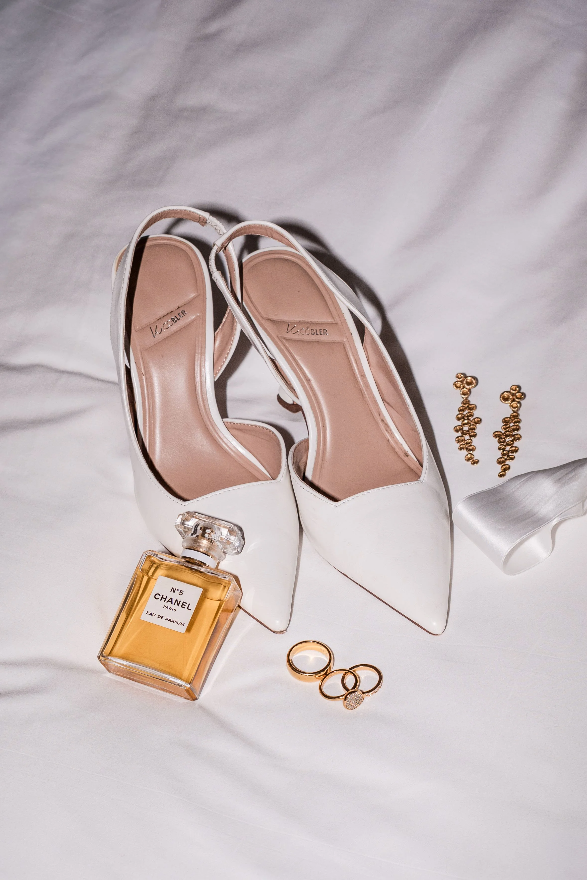 Weiße High Heels, Parfümflasche Chanel No. 5, Goldring, Diamantring, goldene Ohrhänger, weiße Schleife auf weißem Stoff.