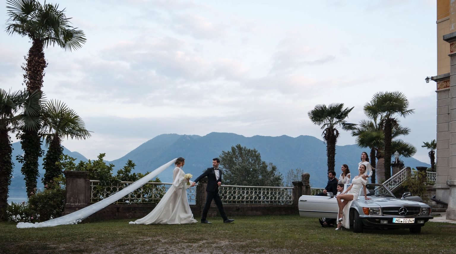 Hochzeit mit Brautpaar, das sich küsst, umgeben von Freunden, Mercedes-Benz Auto, Palmen, Berge im Hintergrund.