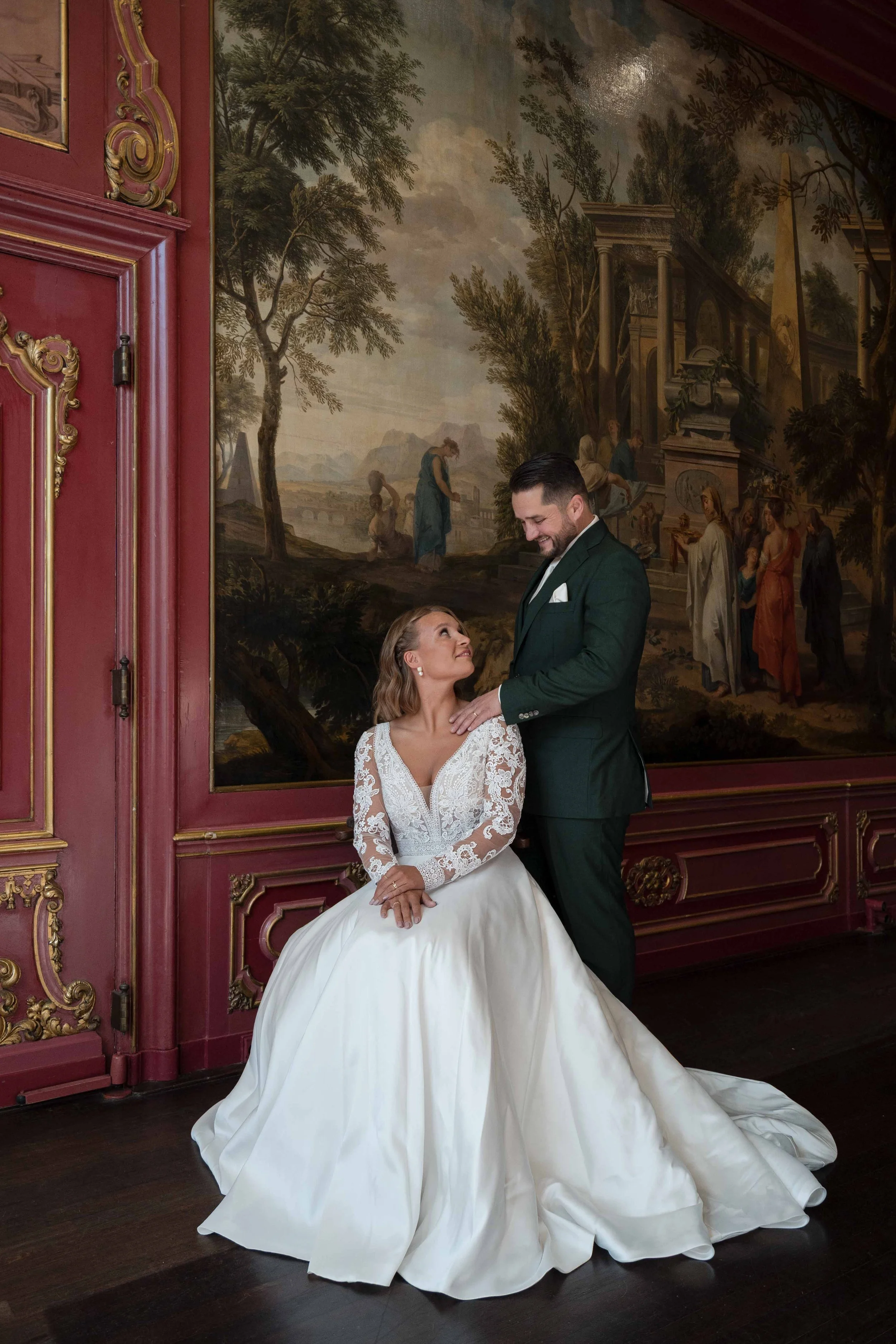 Braut in weißem Hochzeitskleid sitzt auf dem Boden, während der Bräutigam in einem dunklen Anzug steht und sie liebevoll ansieht, vor einer großen Wandmalerei mit Szenen im Grace.