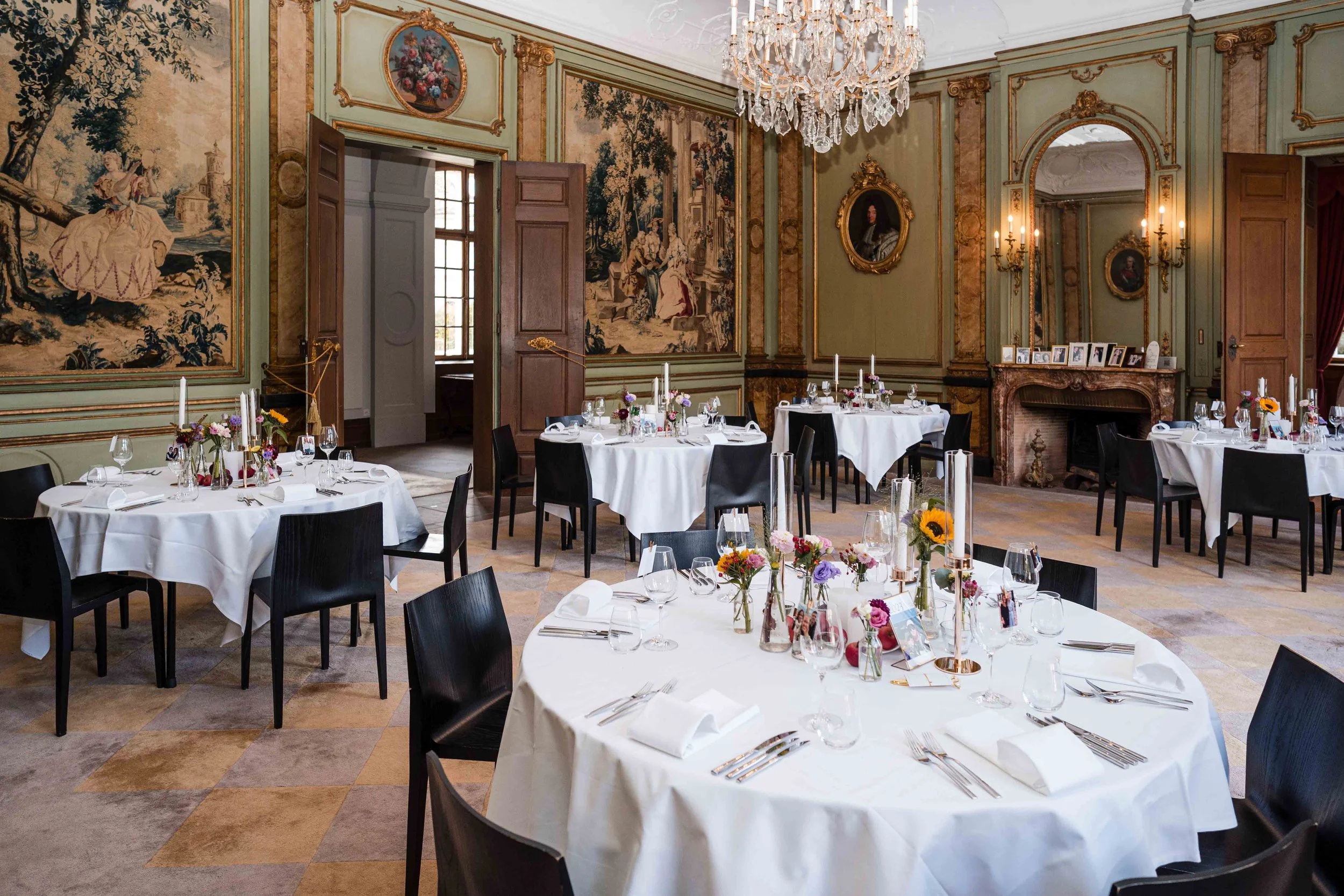 Eleganter Speisesaal mit mehreren runden Tischen, weißer Tischwäsche, Stühlen, Blumenarrangements und Kerzen in einer historischen, kunstvollen Umgebung mit Wandteppichen, Gemälden und Kristalllüstern.
