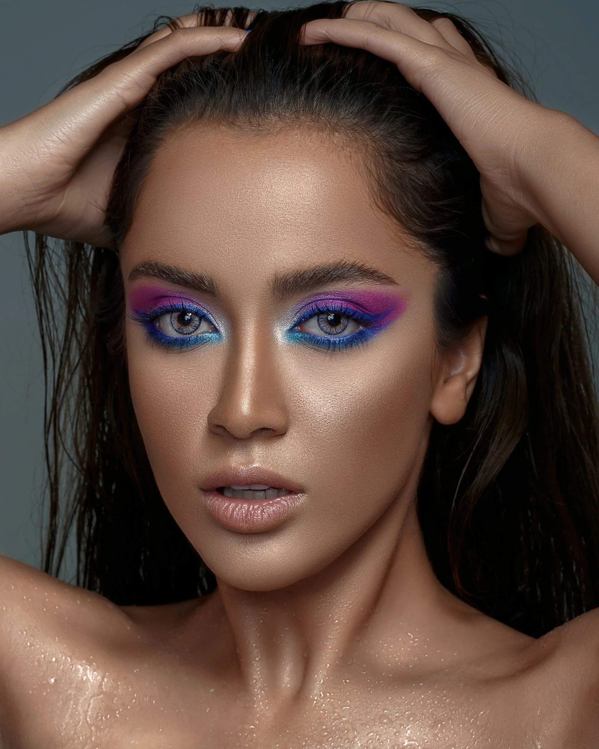 Close-up of a woman with wet skin, holding her hands on her head, showing colorful eye makeup with pink, purple, and blue shades, and water droplets on her shoulders and neck.