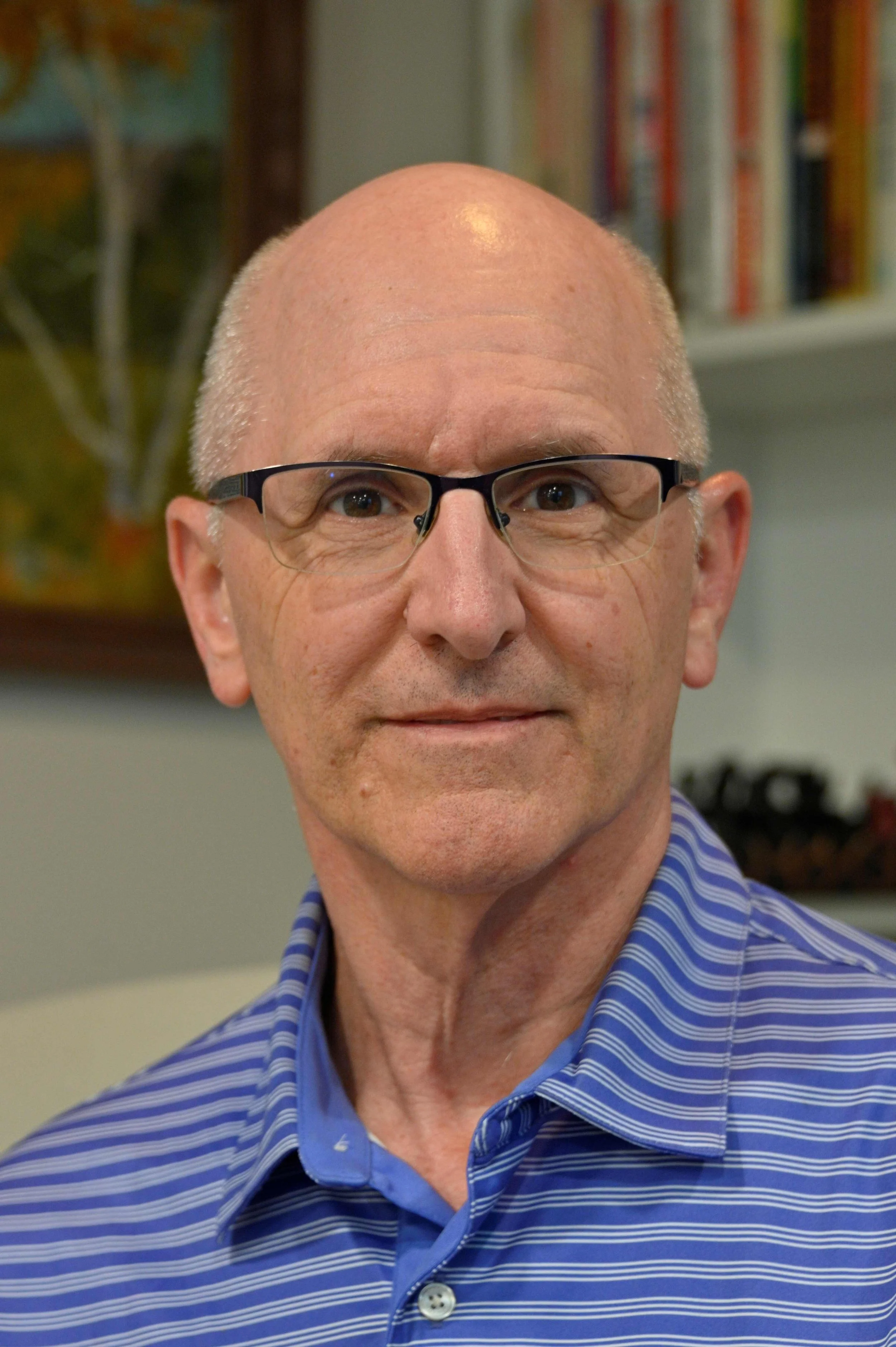 A middle-aged man with glasses and a blue striped dress shirt, smiling slightly, standing in an indoor setting with a bookshelf and a painting in the background.