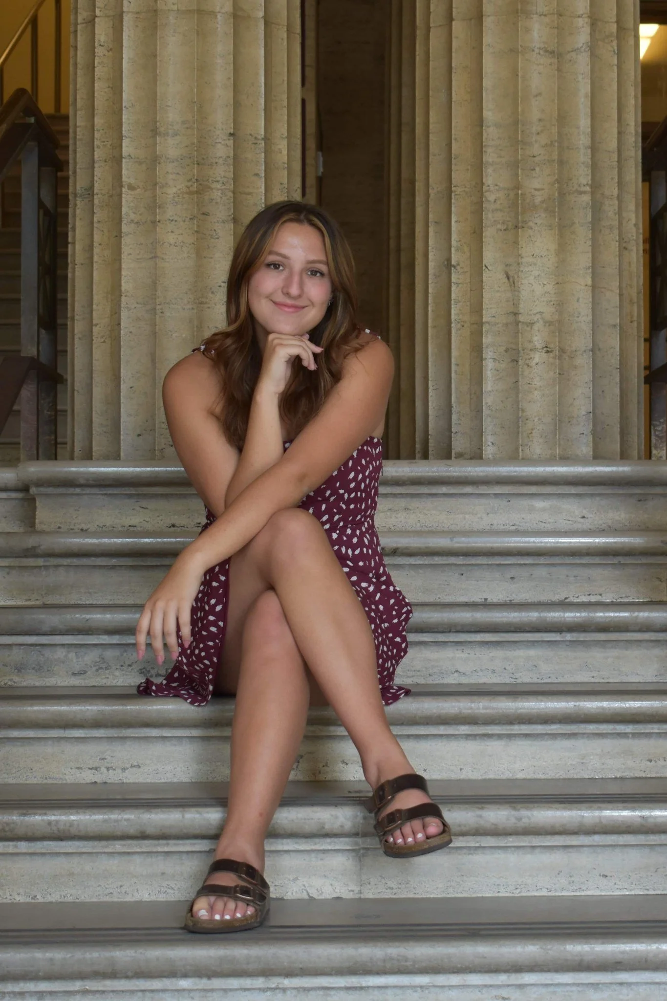 A young woman with long wavy brown hair, sitting on marble stairs in front of beige stone columns. She is wearing a burgundy dress with white polka dots, brown sandals, and is smiling while resting her chin on her hand.