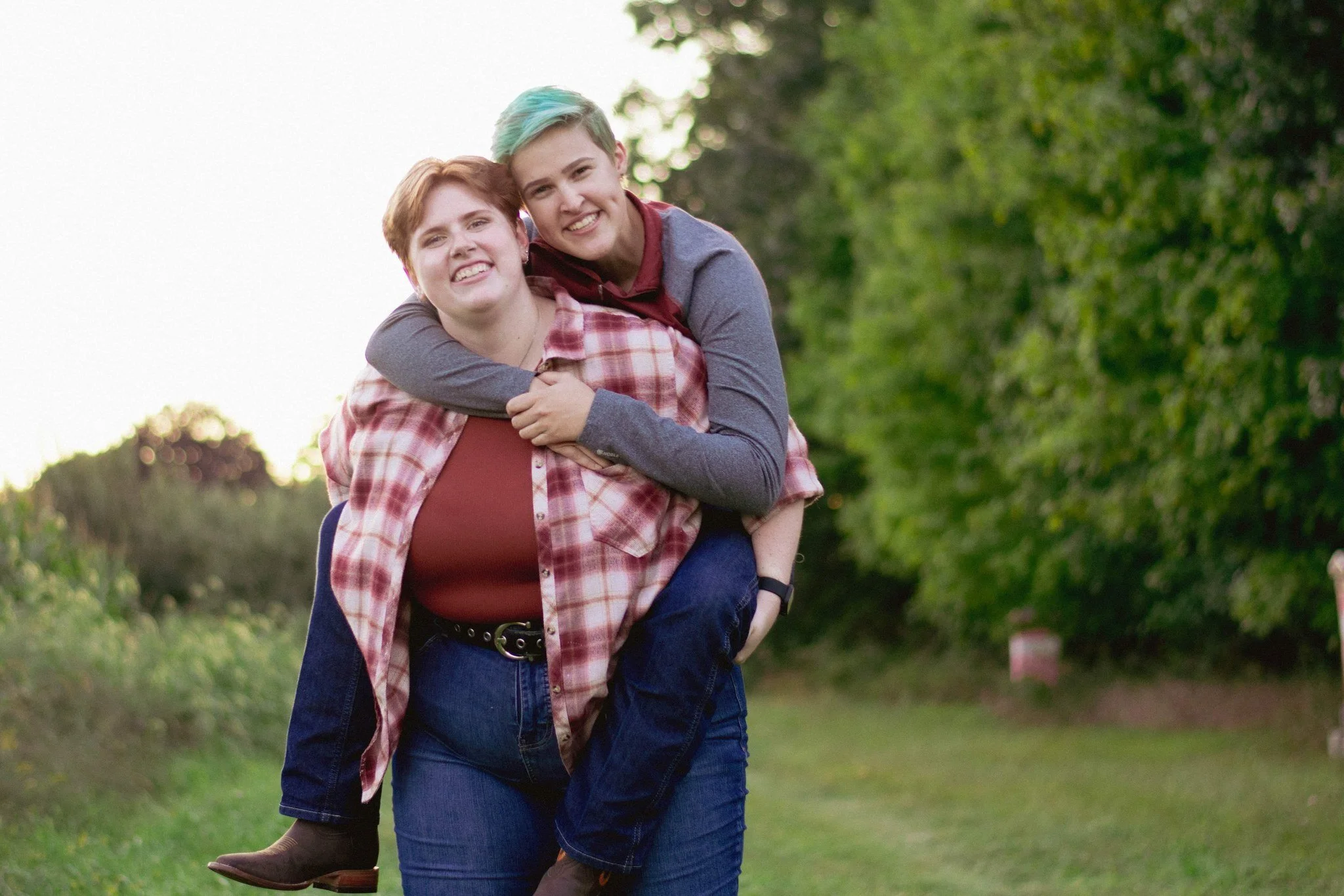 Two young women enjoying a piggyback ride outdoors, smiling, with green trees and grass in the background.