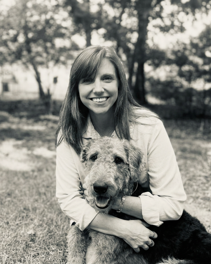 A smiling woman with shoulder-length hair hugging a large, fluffy dog outdoors in a park with trees.