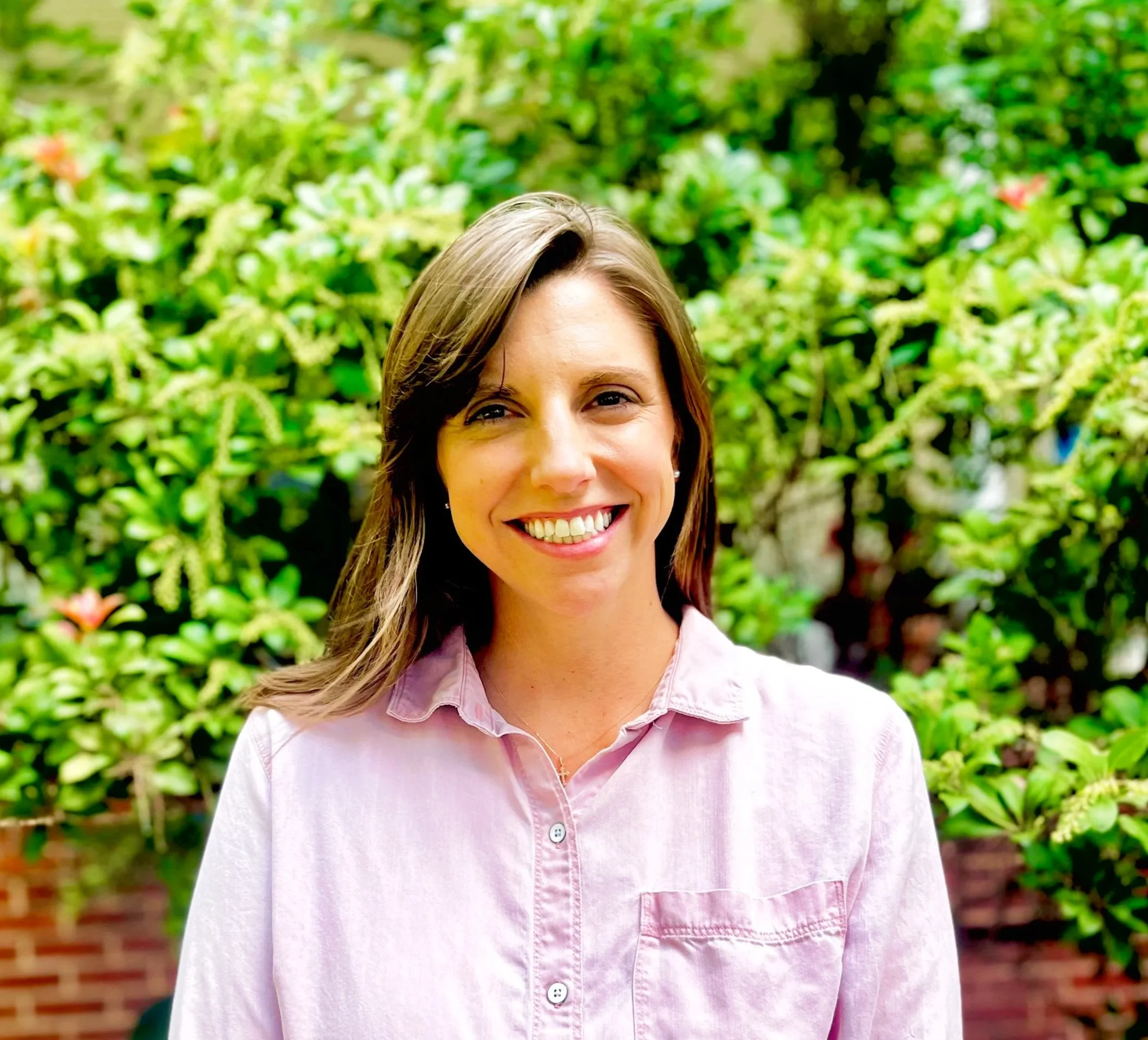 Close-up of a smiling woman with brown hair and light skin, wearing a light pink shirt, outdoors with green foliage background.