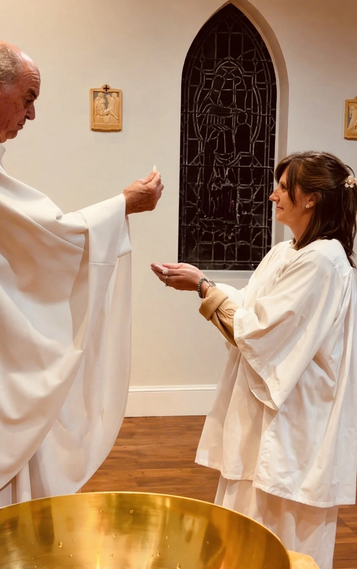 A bishop performing a religious ritual with a woman in a church. The bishop holds a small object, and the woman has her hands opened in a prayer-like position. They both wear white robes. The church has a stained glass window and religious wall art.