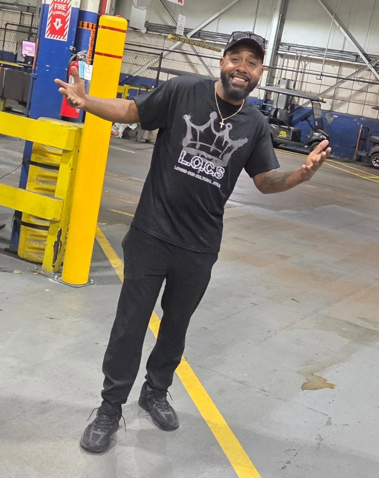 A man standing inside a assembly plant, smiling, wearing a black T-shirt with a Grey crown graphic and the word 'LOCS', black pants, black sneakers, and a black cap, with his arms raised.