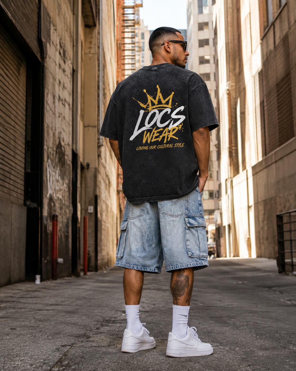 A man standing in an urban alley wearing a black T-shirt with 'LOC'S WEAR' and a crown graphic on the back, light blue cargo shorts, white sneakers, and sunglasses.