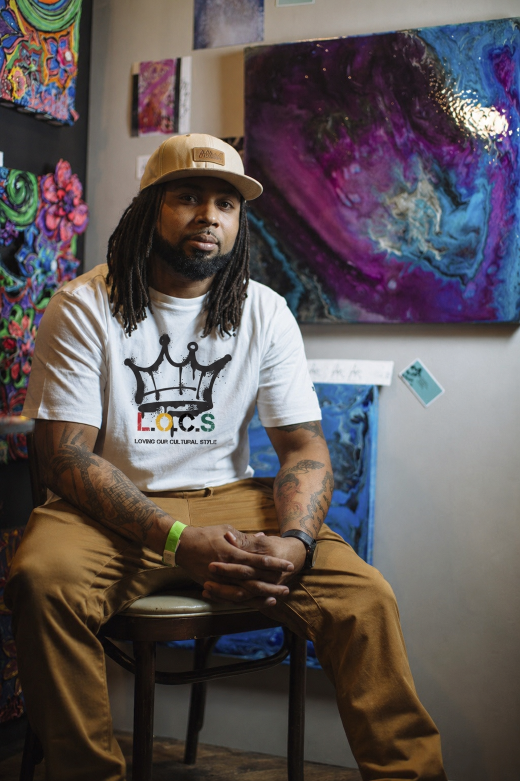 The Owner sitting on a chair in an art gallery, wearing a beige cap and a white T-shirt with a crown graphic and the text 'L.O.C.S' and 'Loving Our Cultural Style', with colorful abstract paintings in the background.